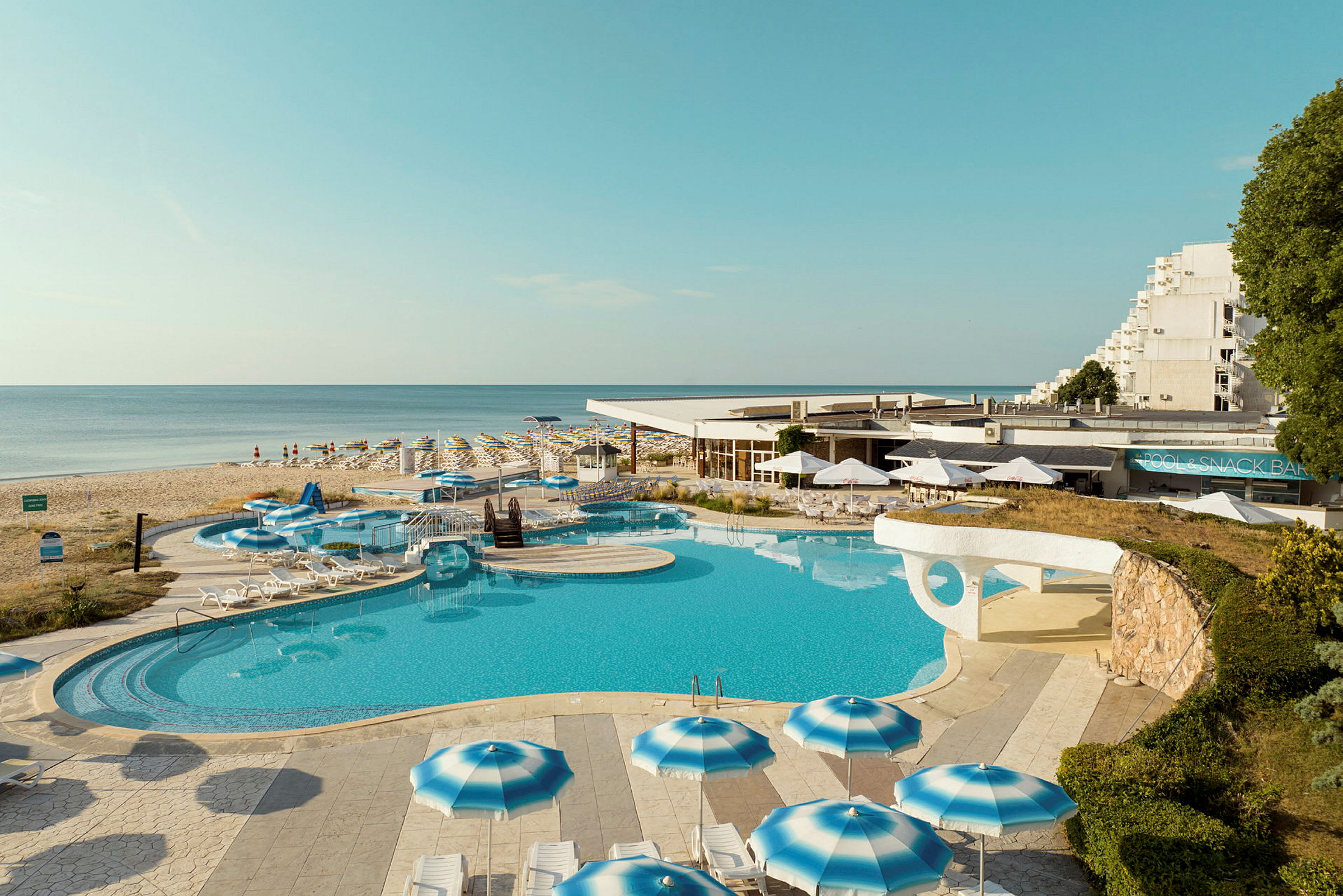 Hotell med poolområde och parasoller vid stranden. (AI-genererad text)