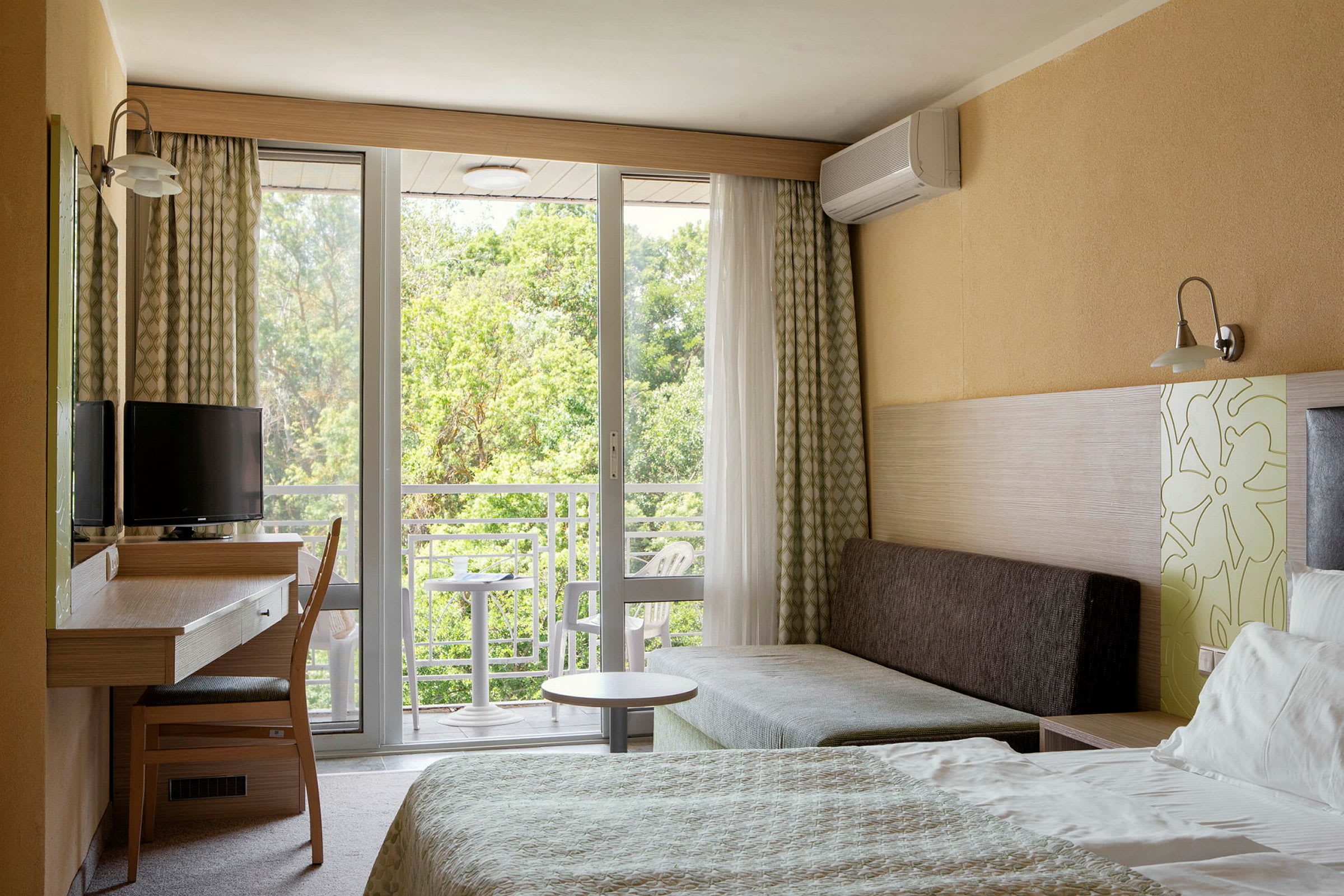 Ett hotellrum med säng, skrivbord, stol och soffa. Fönster och balkong med utsikt mot grönska. (AI-genererad text)
