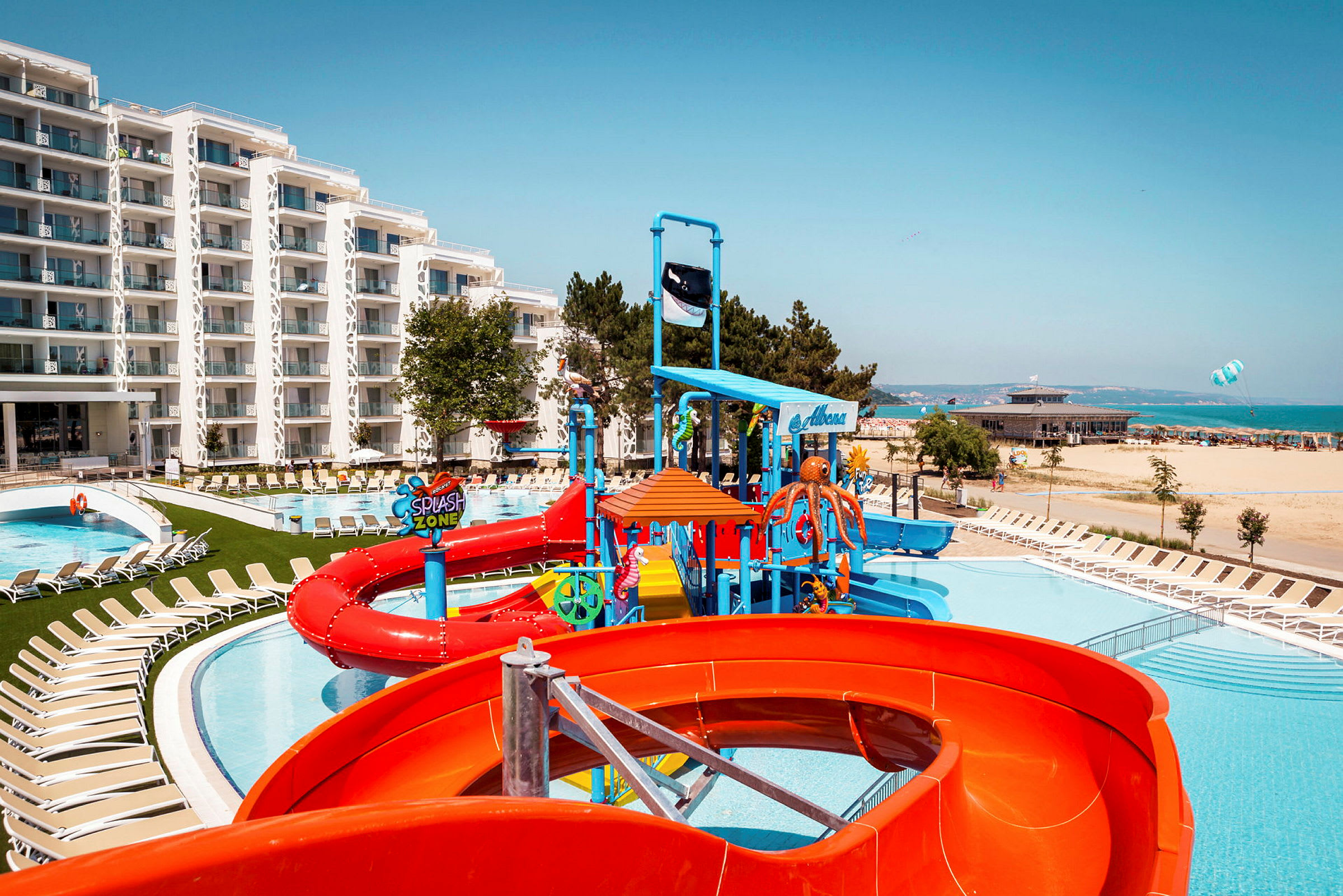 Bild av ett hotellområde med pool, vattenrutschbanor och liggstolar nära stranden. (AI-genererad text)