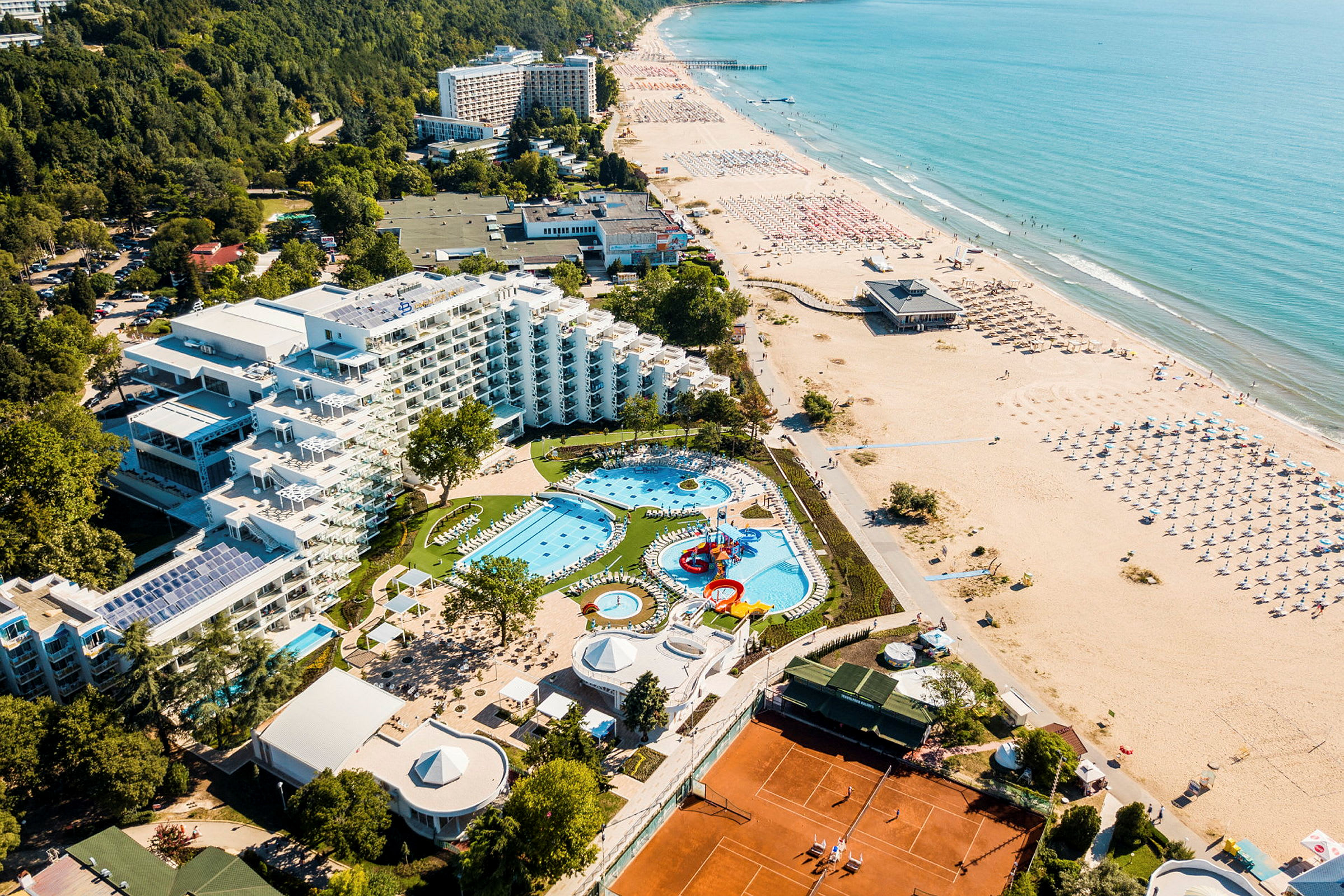 Flygbild över hotell med poolområde, grönska och strand. (AI-genererad text)