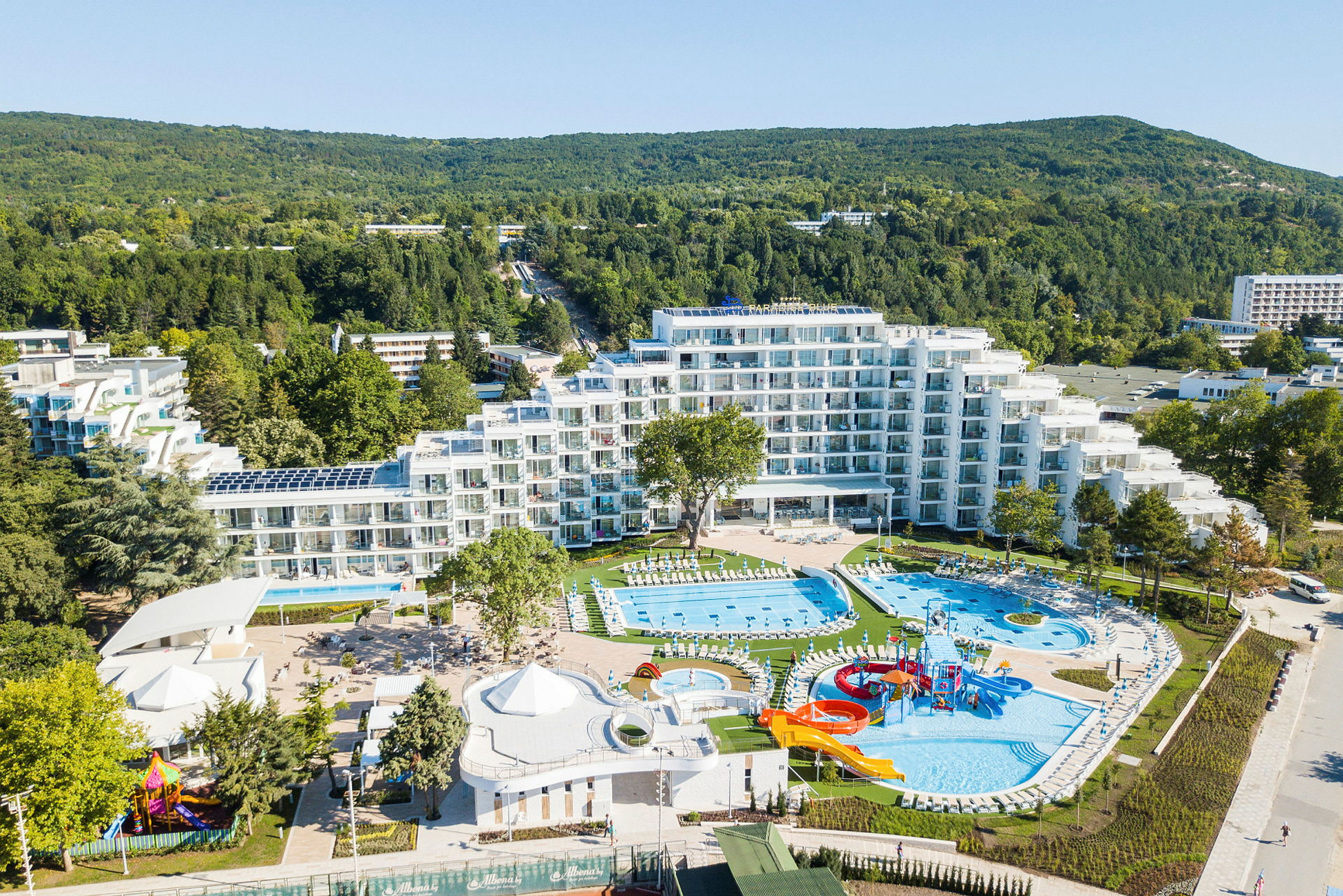 Flygfoto över ett hotell med pooler och lekplatser, omgiven av grönska och mindre byggnader. (AI-genererad text)