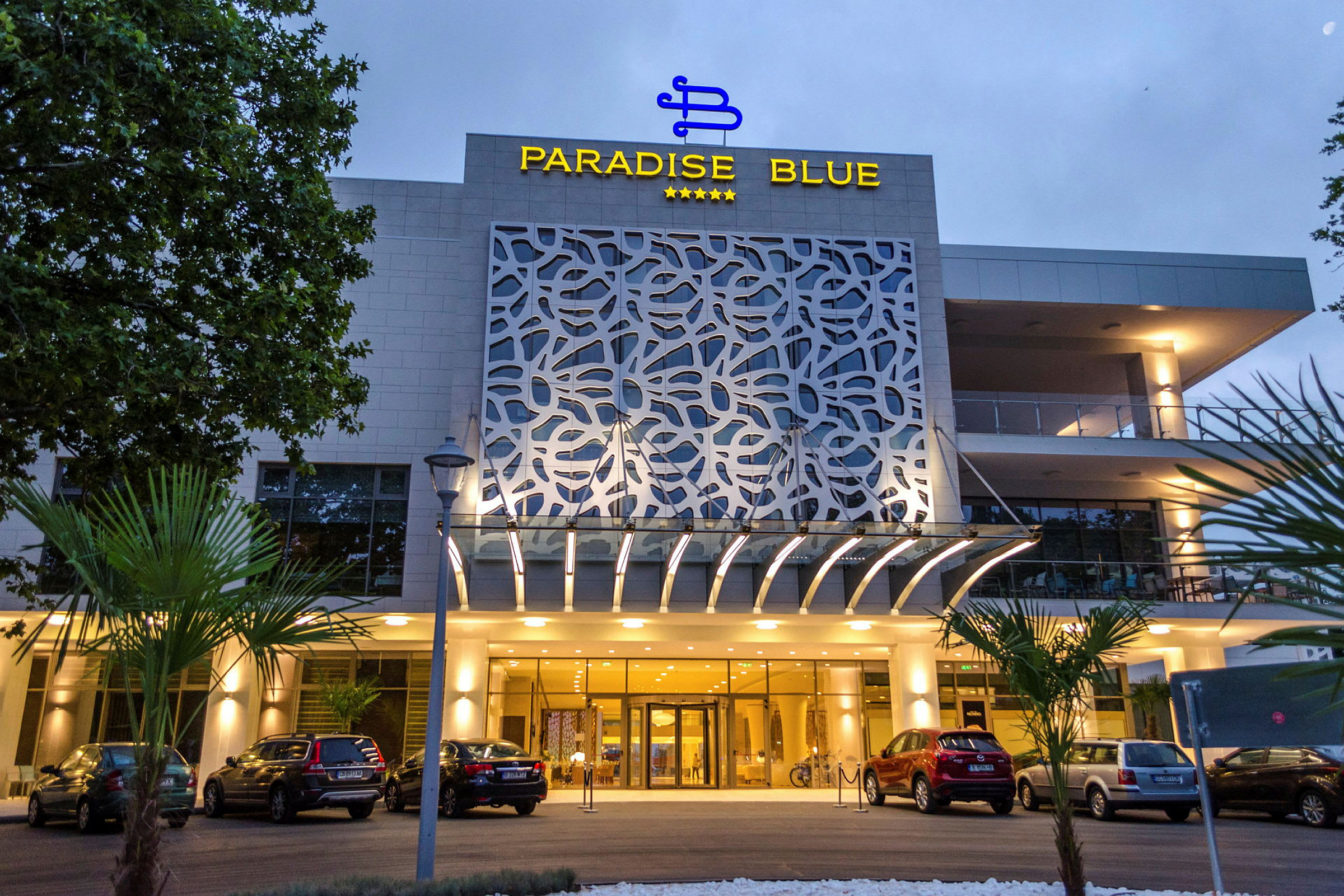 Fasaden på Paradise Blue hotell med bilparkering och entré. (AI-genererad text)