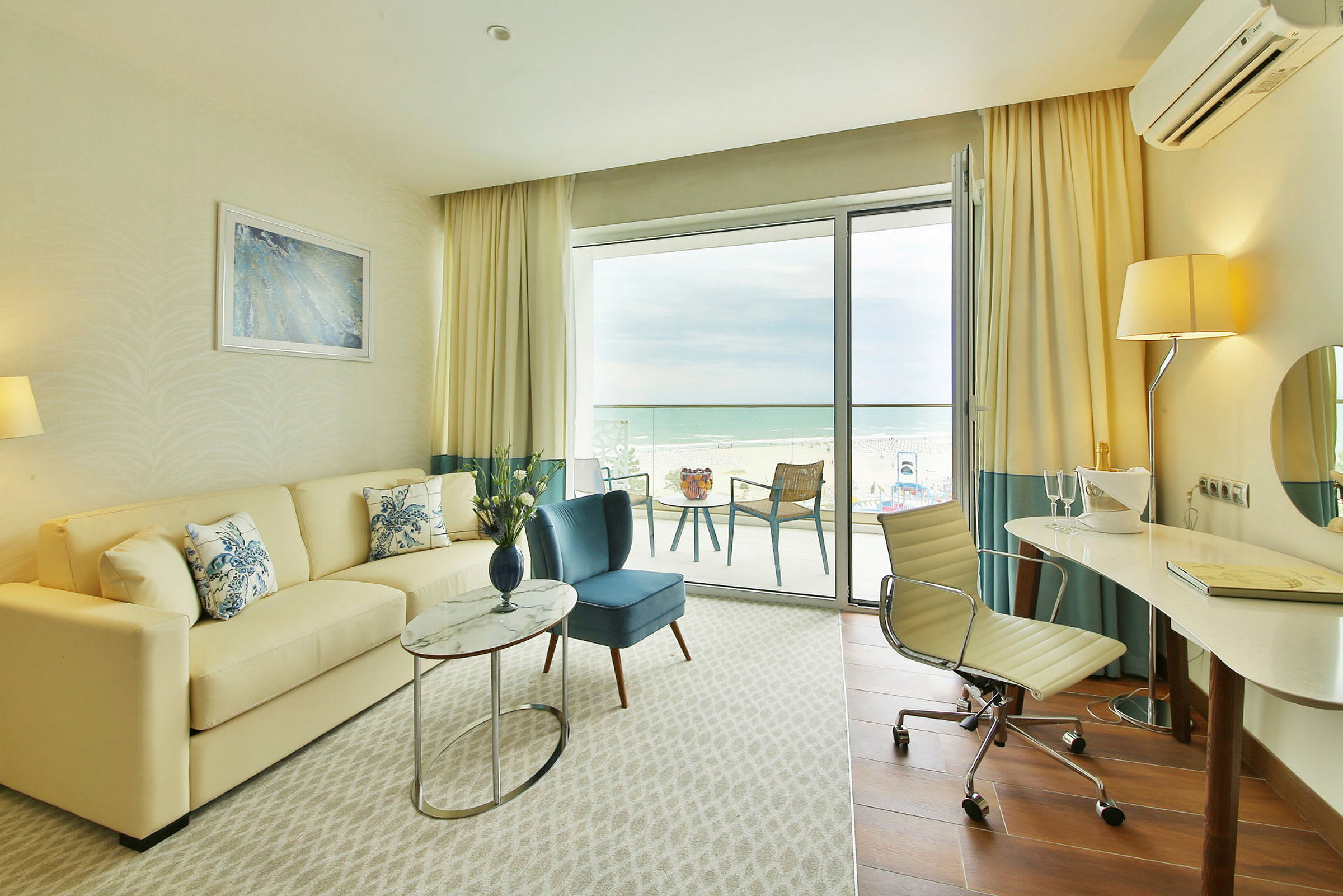 Ett ljust hotellrum med en soffa, fåtölj, ett skrivbord och en balkong med utsikt över stranden. (AI-genererad text)