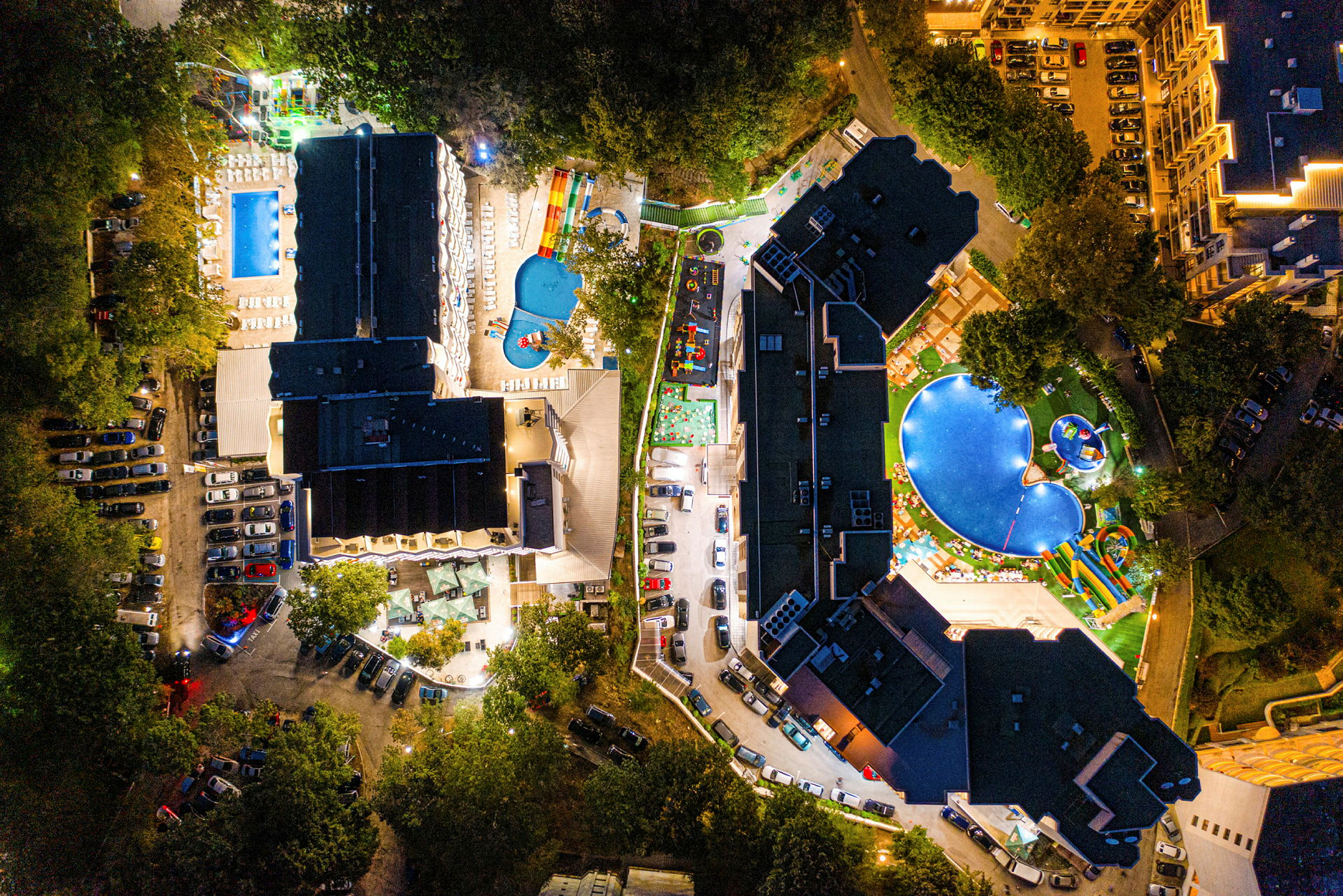 Flygfoto av hotellområde med pooler, byggnader och parkeringsplatser, upplyst på kvällen. (AI-genererad text)