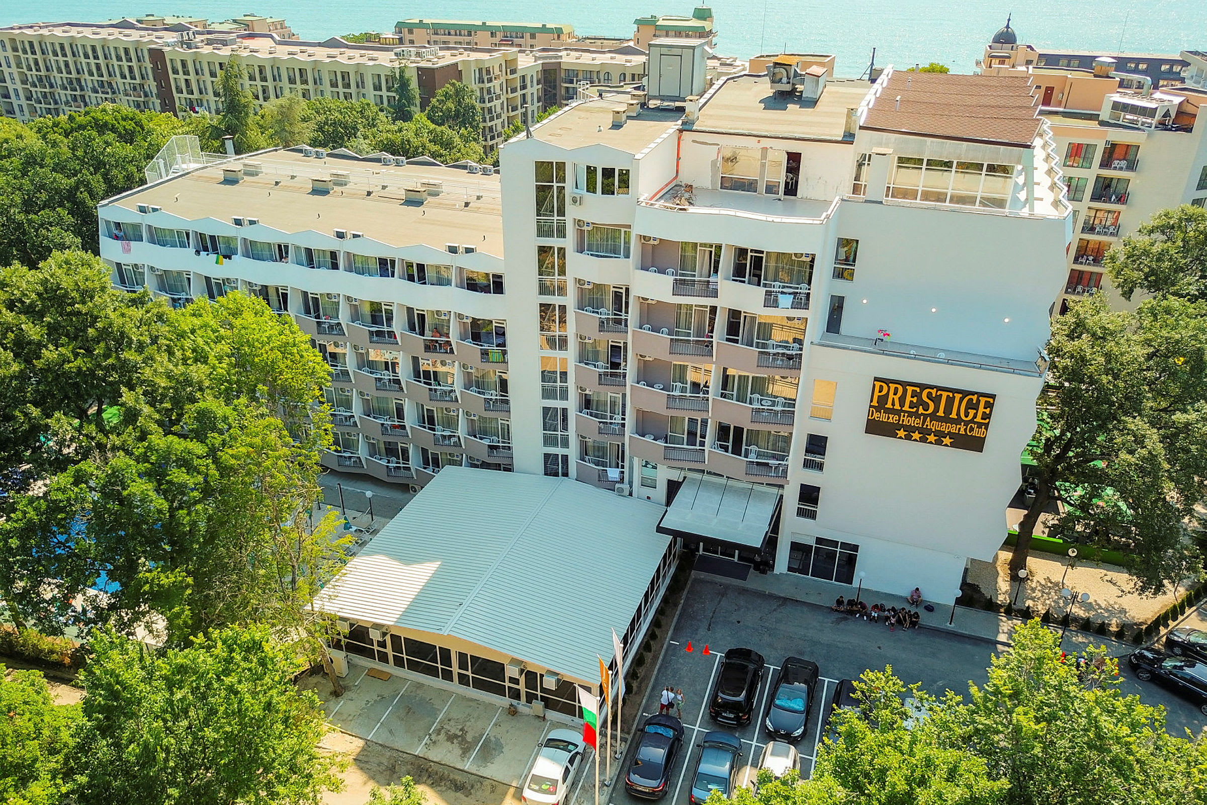 Bild av ett hotellkomplex med flerbostadshus och balkonger, omgiven av grönska och med parkerade bilar framför entrén. (AI-genererad text)