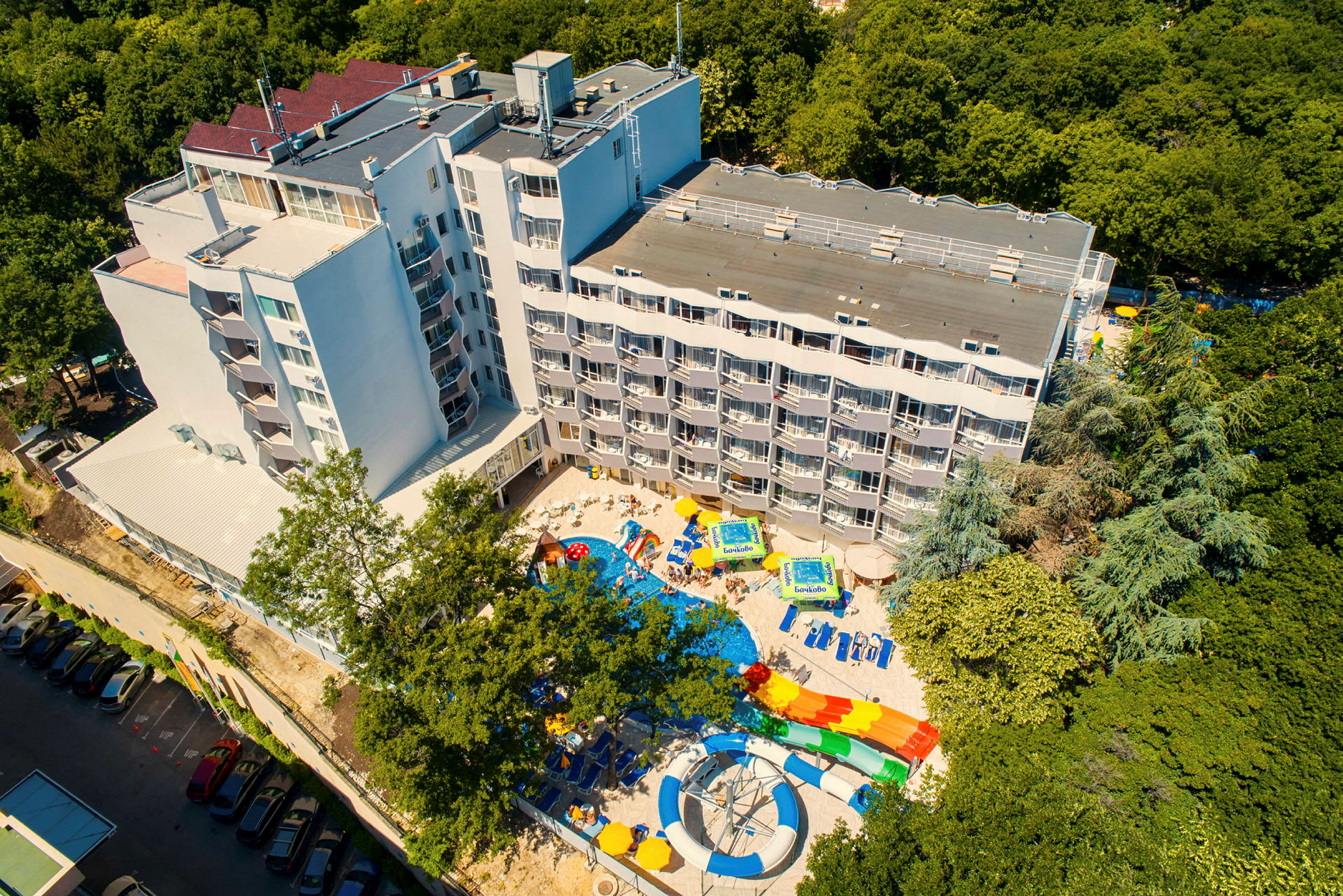 Ett hotell med flera våningar, omgiven av grönska. En pool med vattenrutschbanor och solstolar finns framför byggnaden. (AI-genererad text)