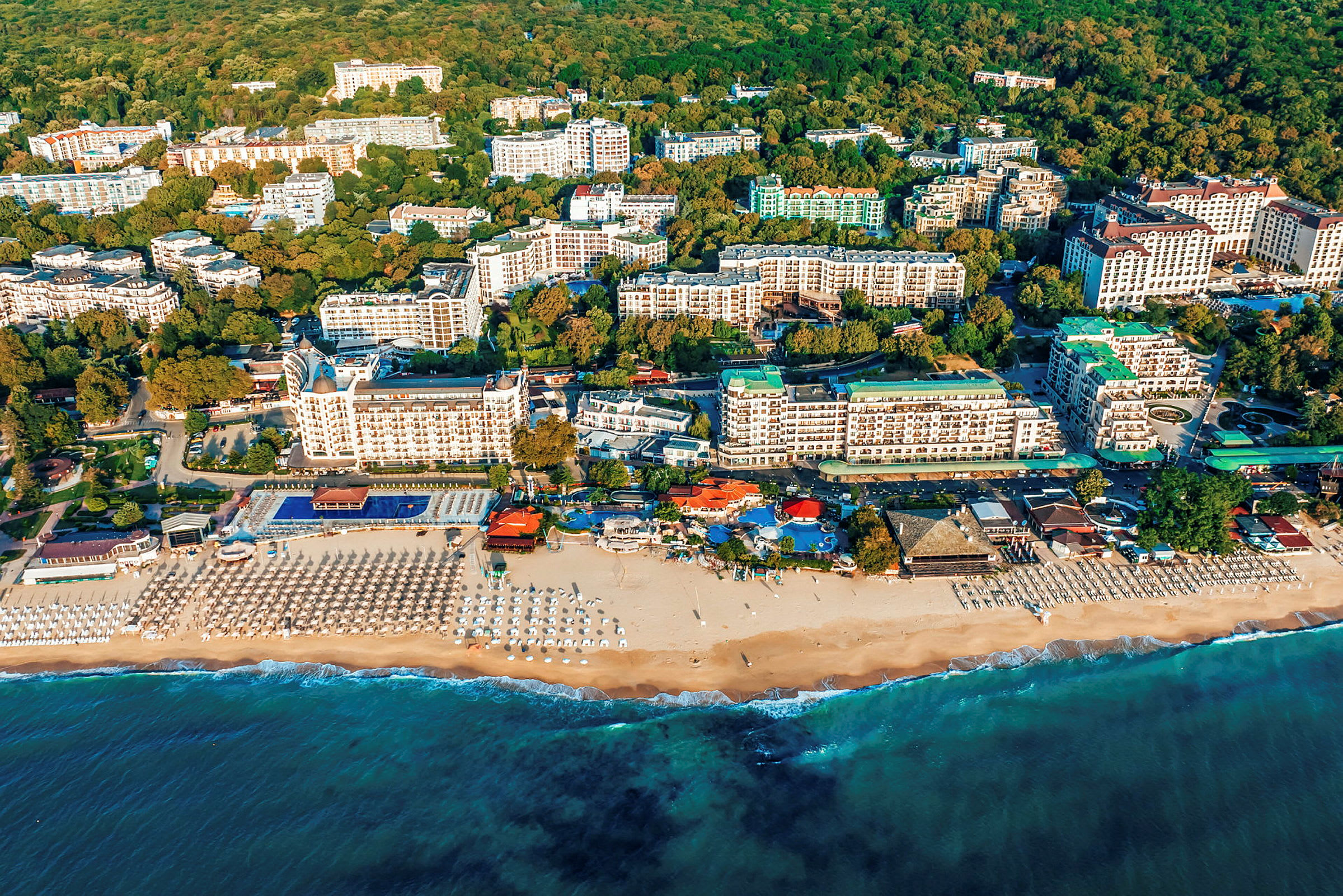 Flygfoto över en kuststad med stranden, havet och flera hotellbyggnader som omges av grönska. (AI-genererad text)