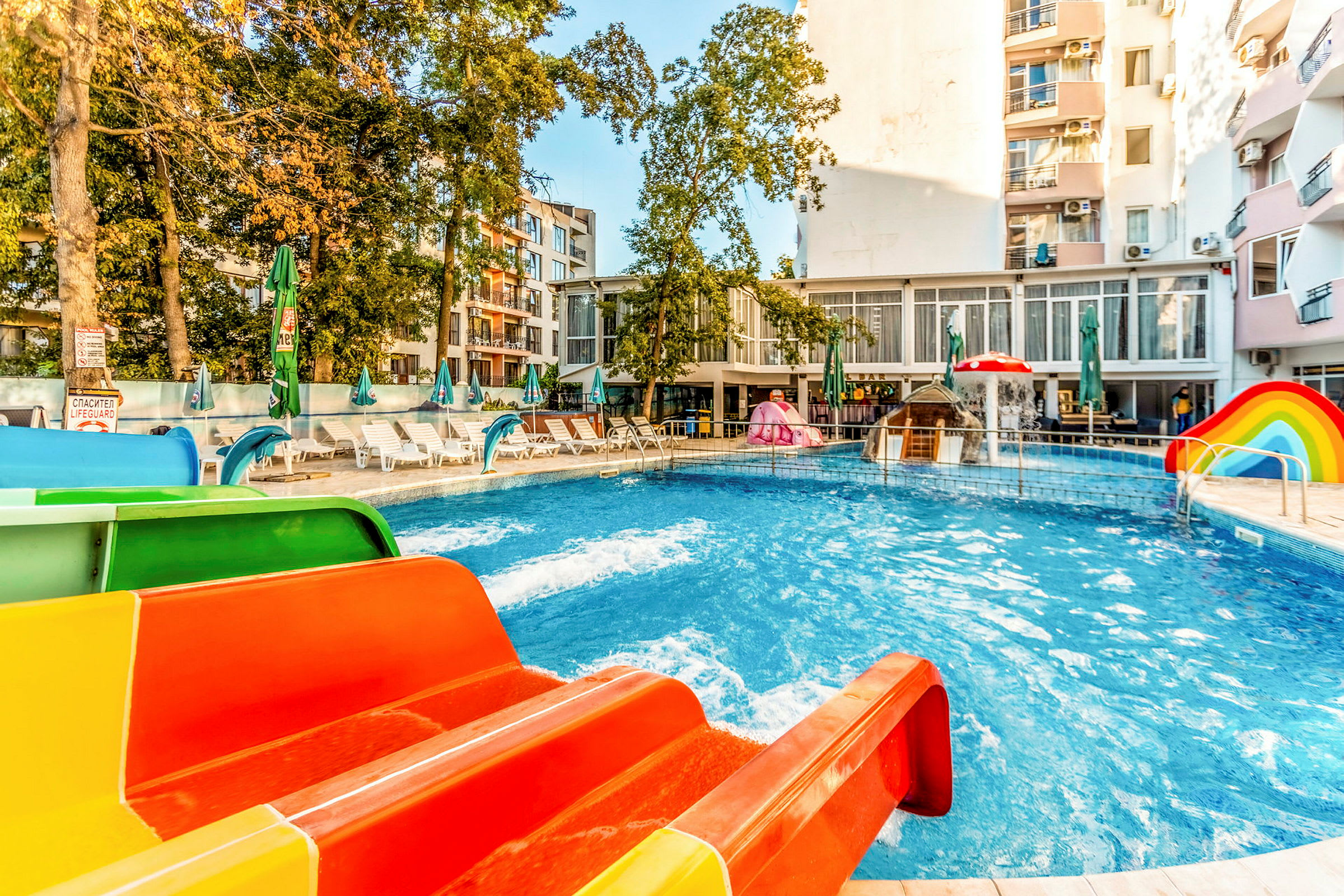 En swimmingpool med färgglada vattenrutschbanor och solstolar runtom, omgiven av byggnader och träd. (AI-genererad text)