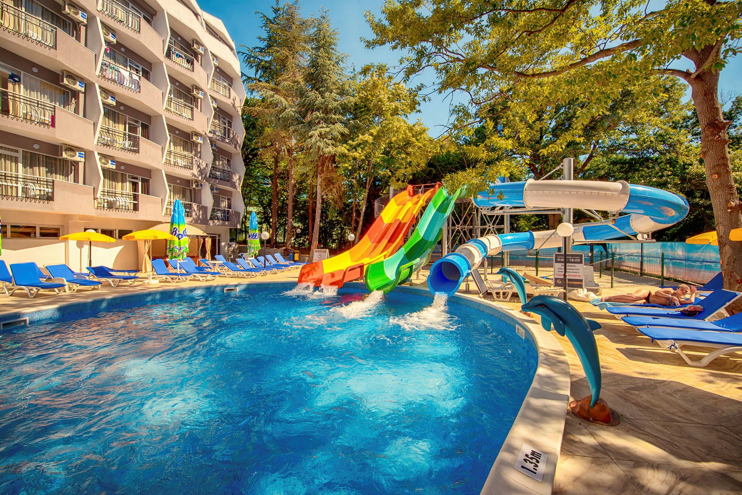 En utomhuspool med färgglada vattenrutschbanor och solstolar runt omkring. Ett hotell med balkonger syns i bakgrunden. (AI-genererad text)