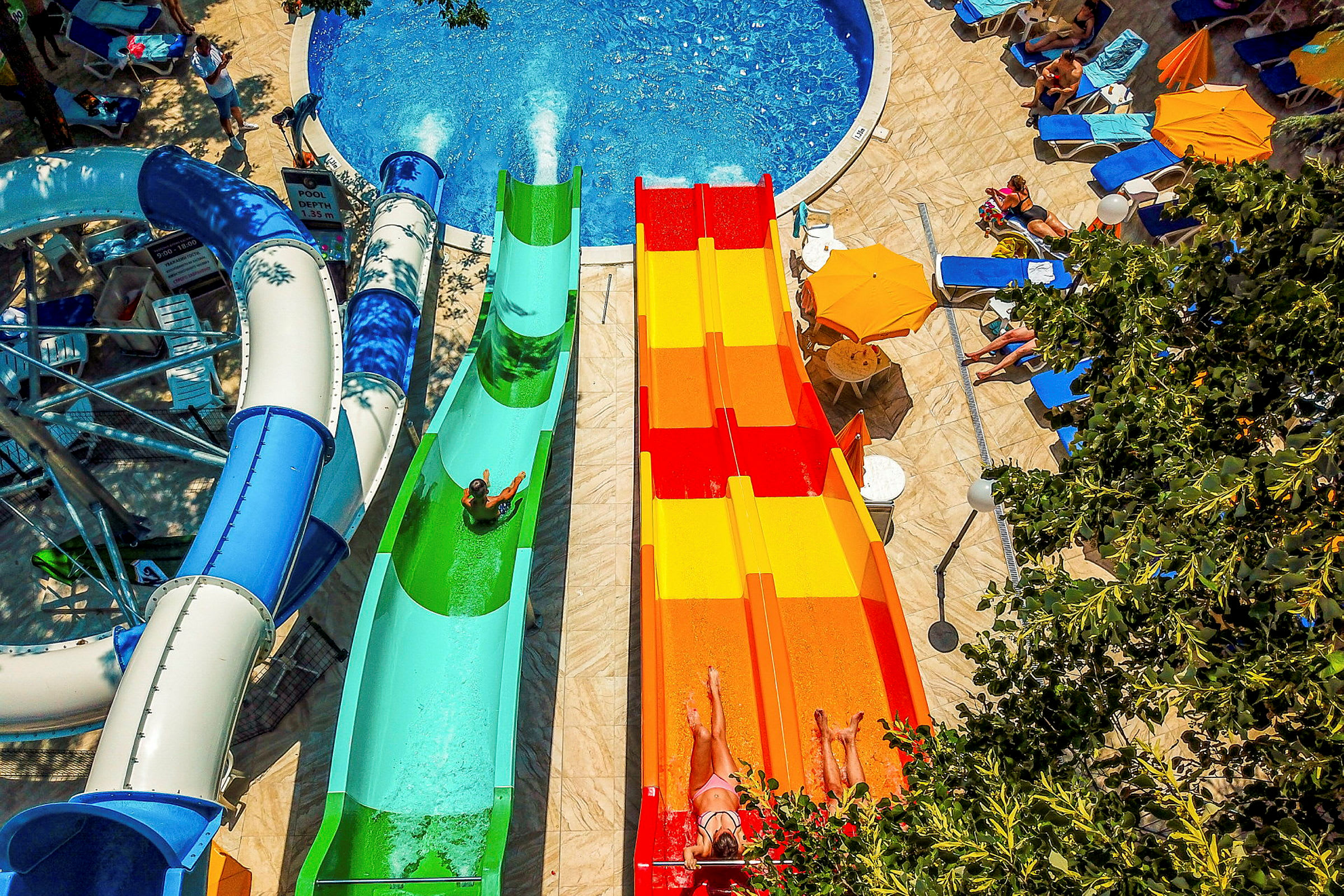 Bilden visar en pool med flera vattenrutschbanor i olika färger. Det finns människor som åker i rutschbanorna samt solstolar och parasoller runt omkring. (AI-genererad text)