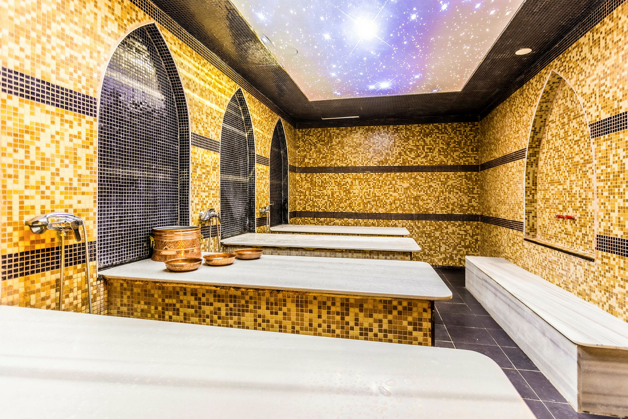 Bild av en hamam med mosaikbeklädda väggar, vita marmorbänkar och kopparkärl. (AI-genererad text)