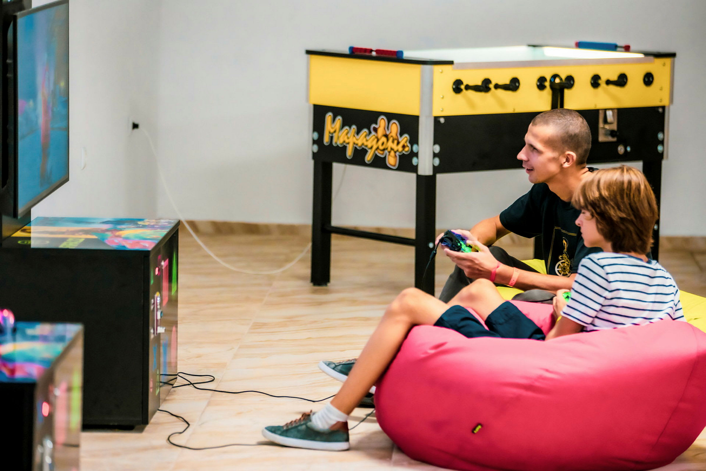 Två personer sitter i en färgad sittsäckssoffa och spelar tv-spel framför en skärm, med ett bordshockeyspel i bakgrunden. (AI-genererad text)