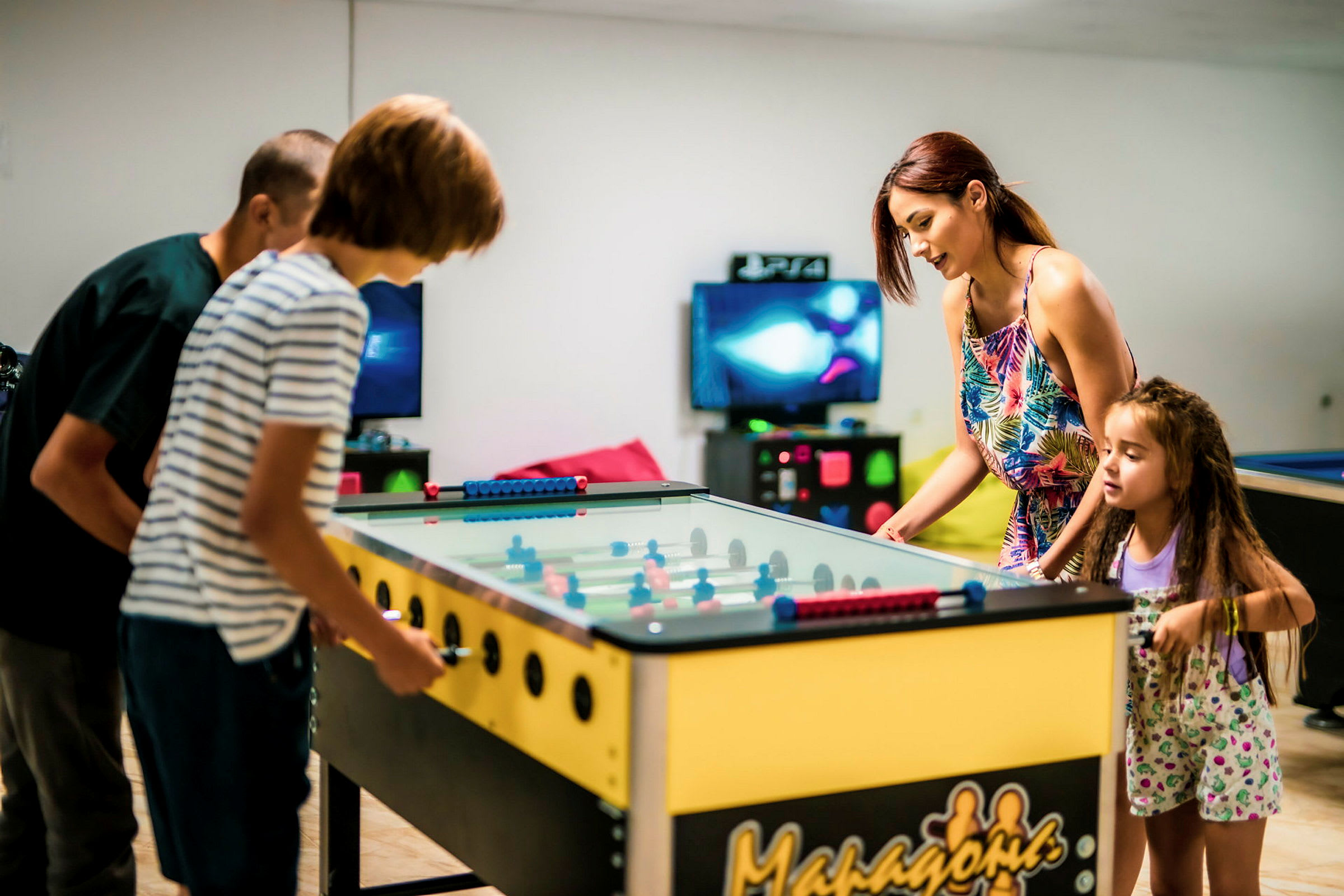 Fyra personer spelar foosball på ett bord i ett rum med spelkonsoler i bakgrunden. (AI-genererad text)
