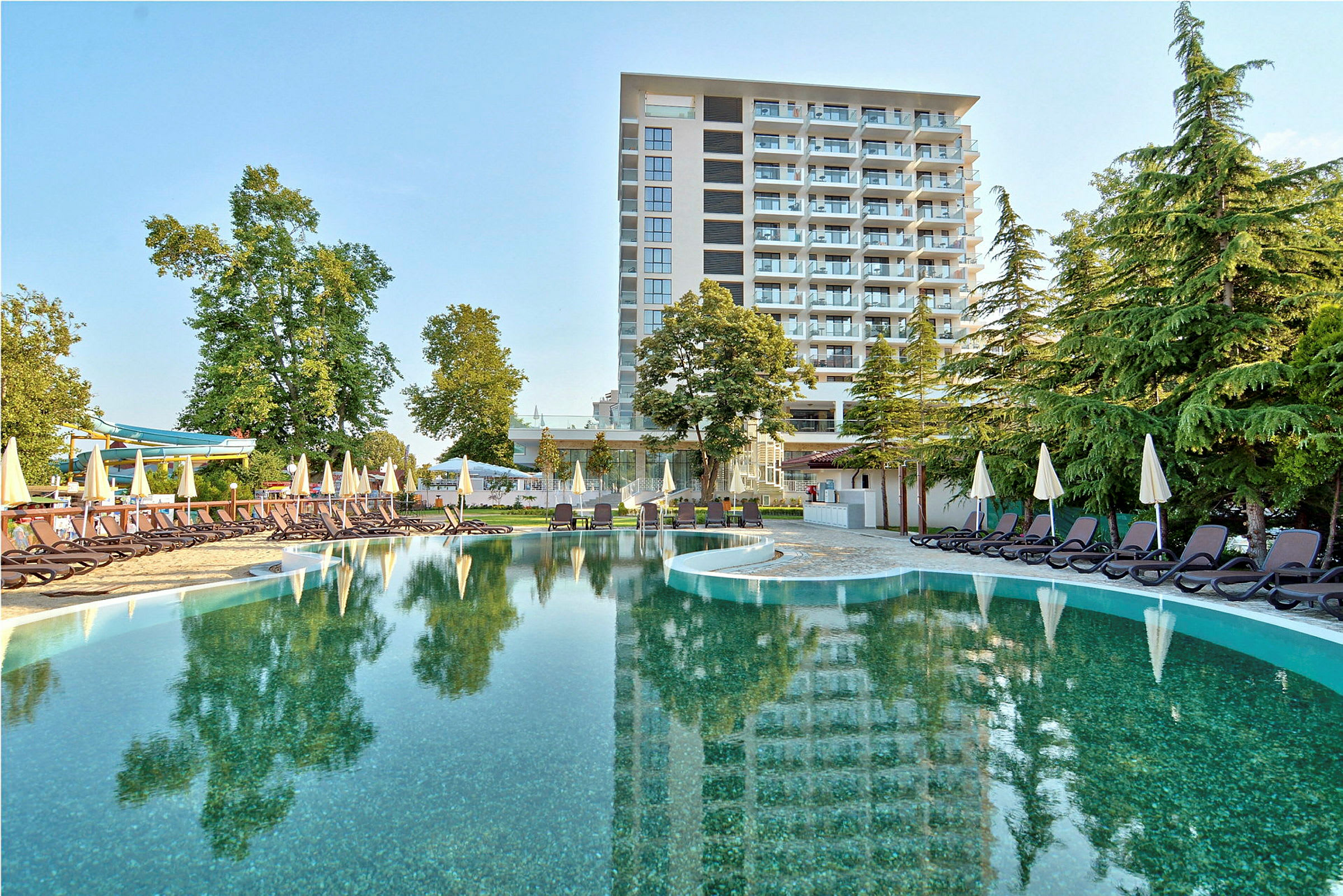 Hotell med turkost vatten vid poolområdet, omgiven av solbäddar och gröna träd. (AI-genererad text)