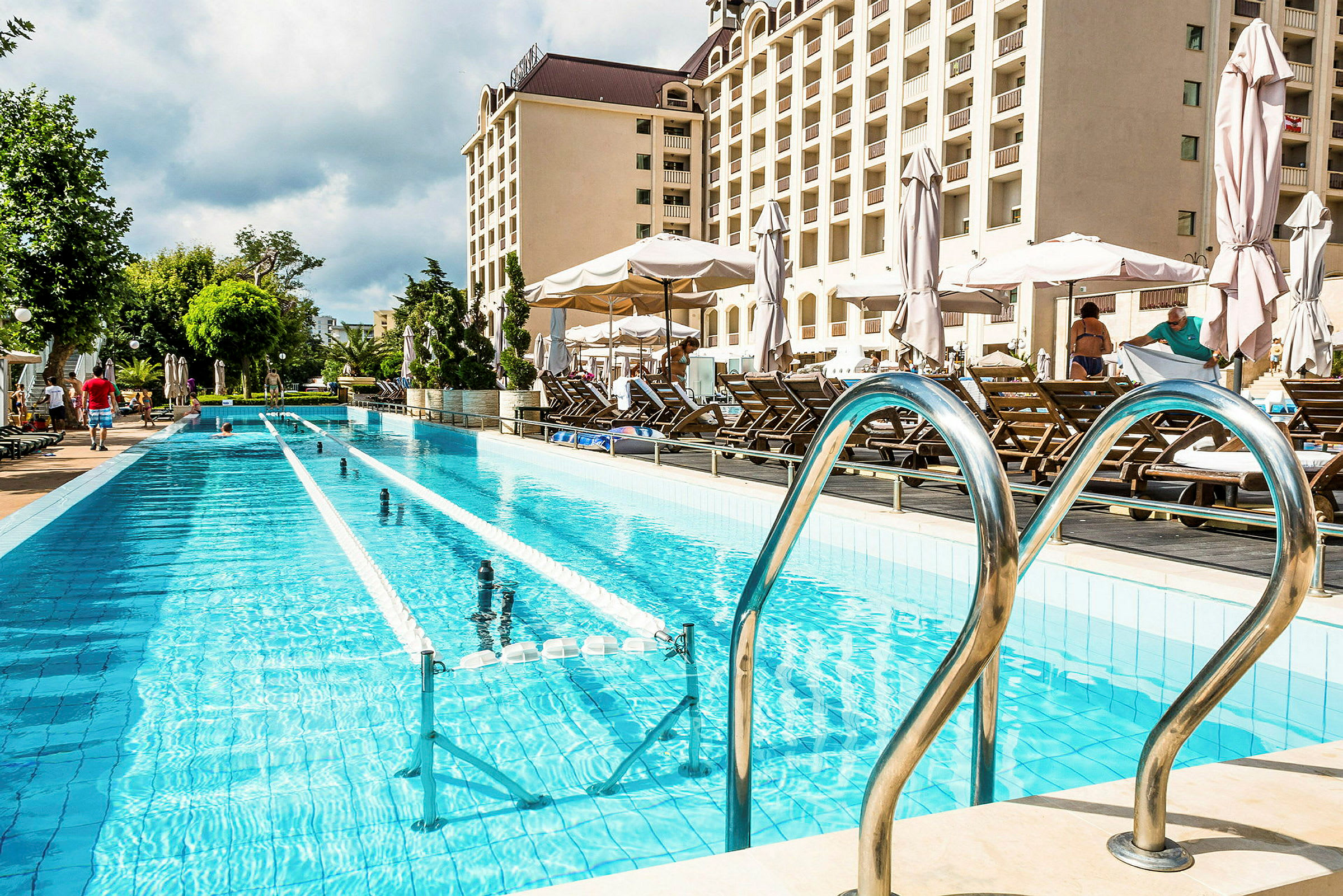 Lång pool med solstolar och parasoller bredvid, hotellbyggnad i bakgrunden. (AI-genererad text)