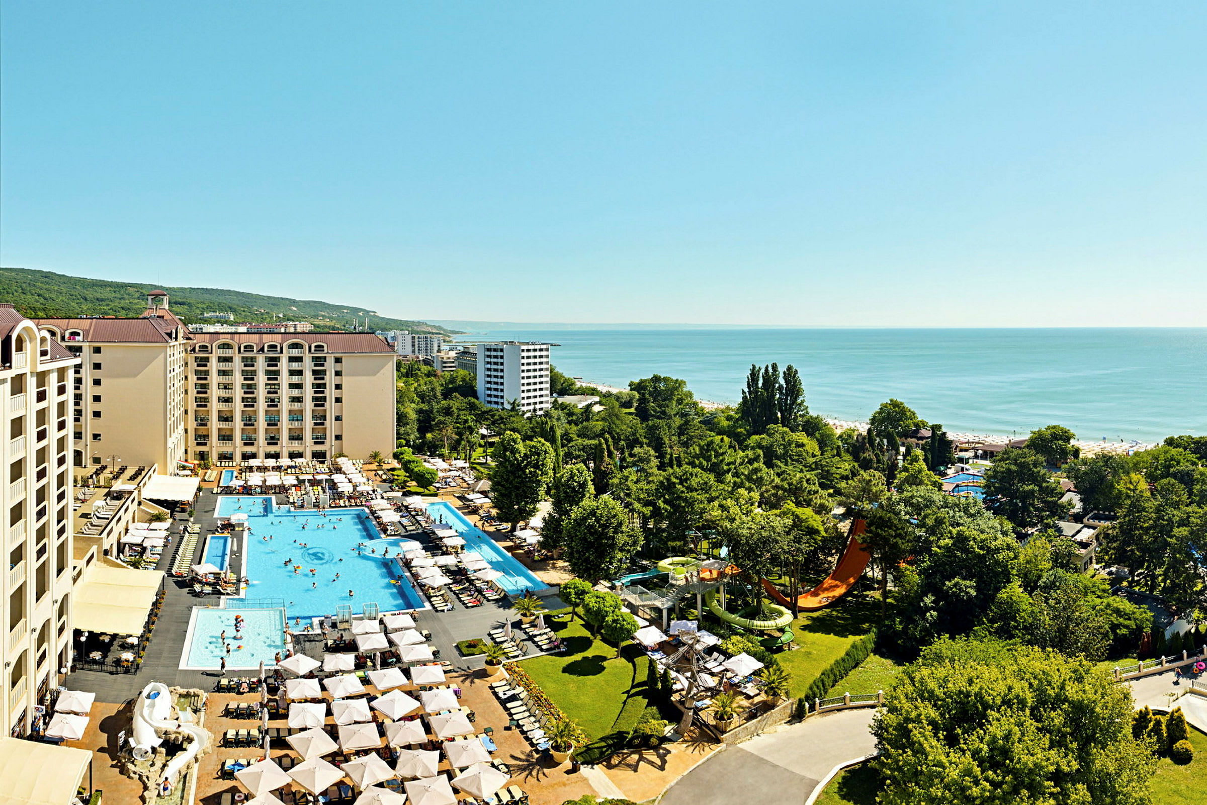 Flygbild av hotellkomplex med pooler, solstolar och grönska, beläget nära havet. (AI-genererad text)