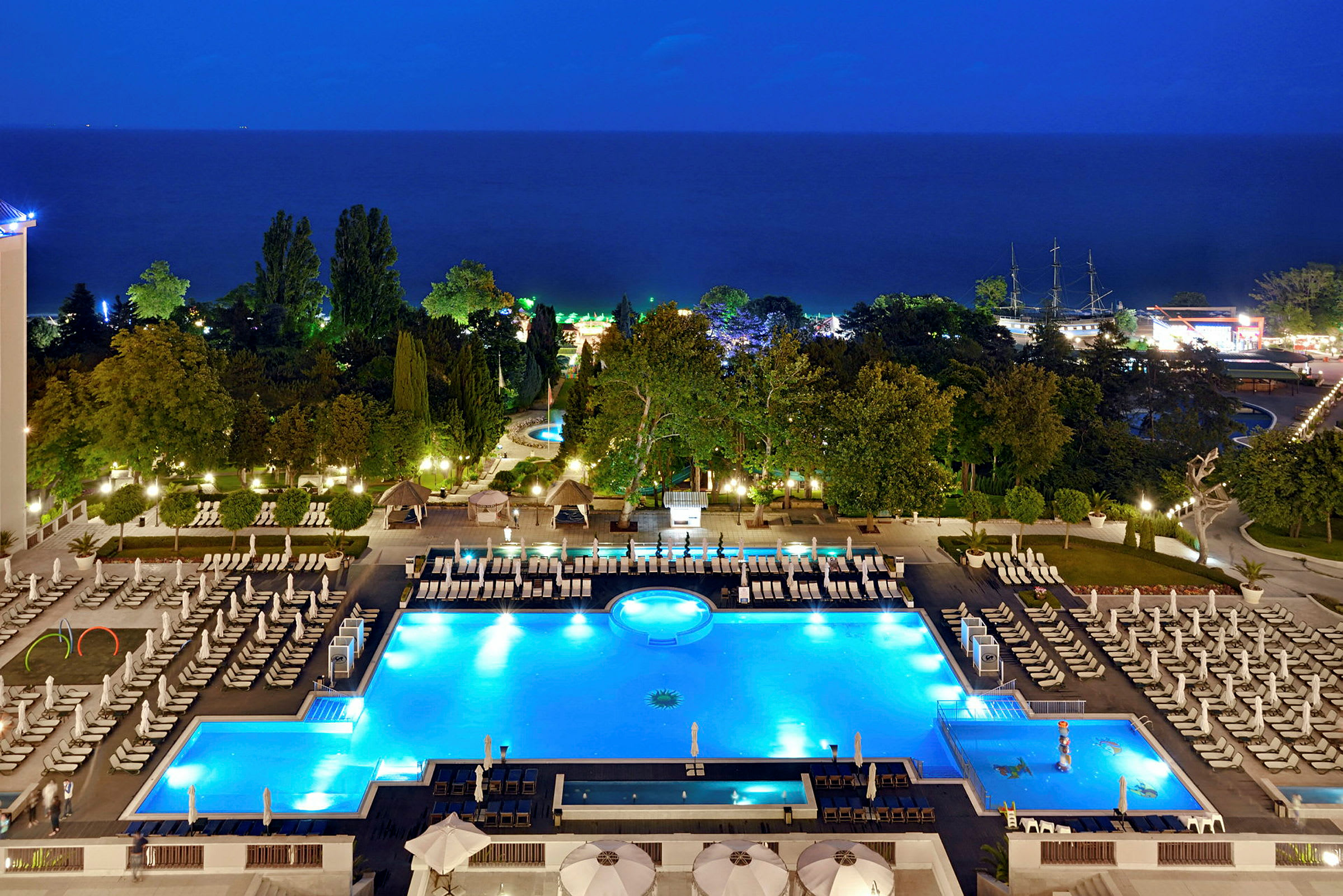 Hotellområde med upplysta pooler och solstolar på kvällen, med träd och hav i bakgrunden. (AI-genererad text)