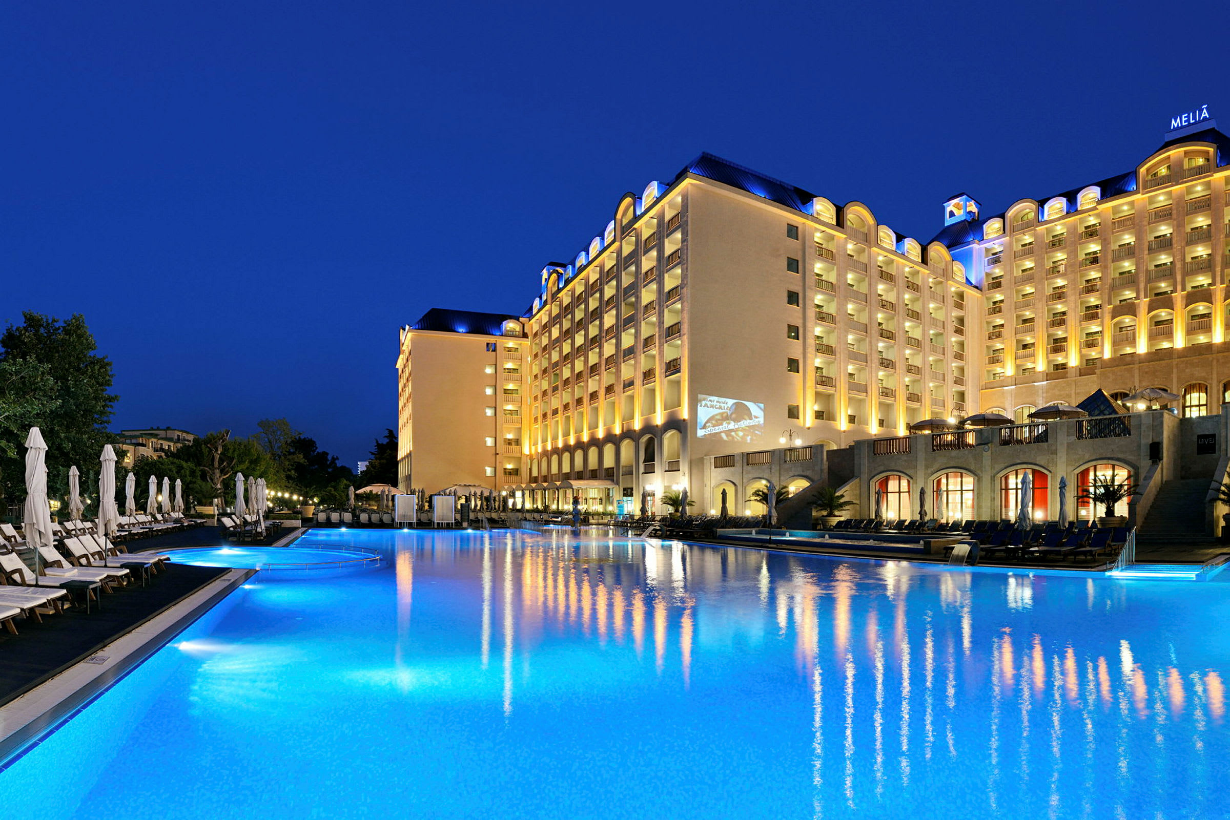 Hotellbyggnad vid en upplyst pool på kvällen. (AI-genererad text)