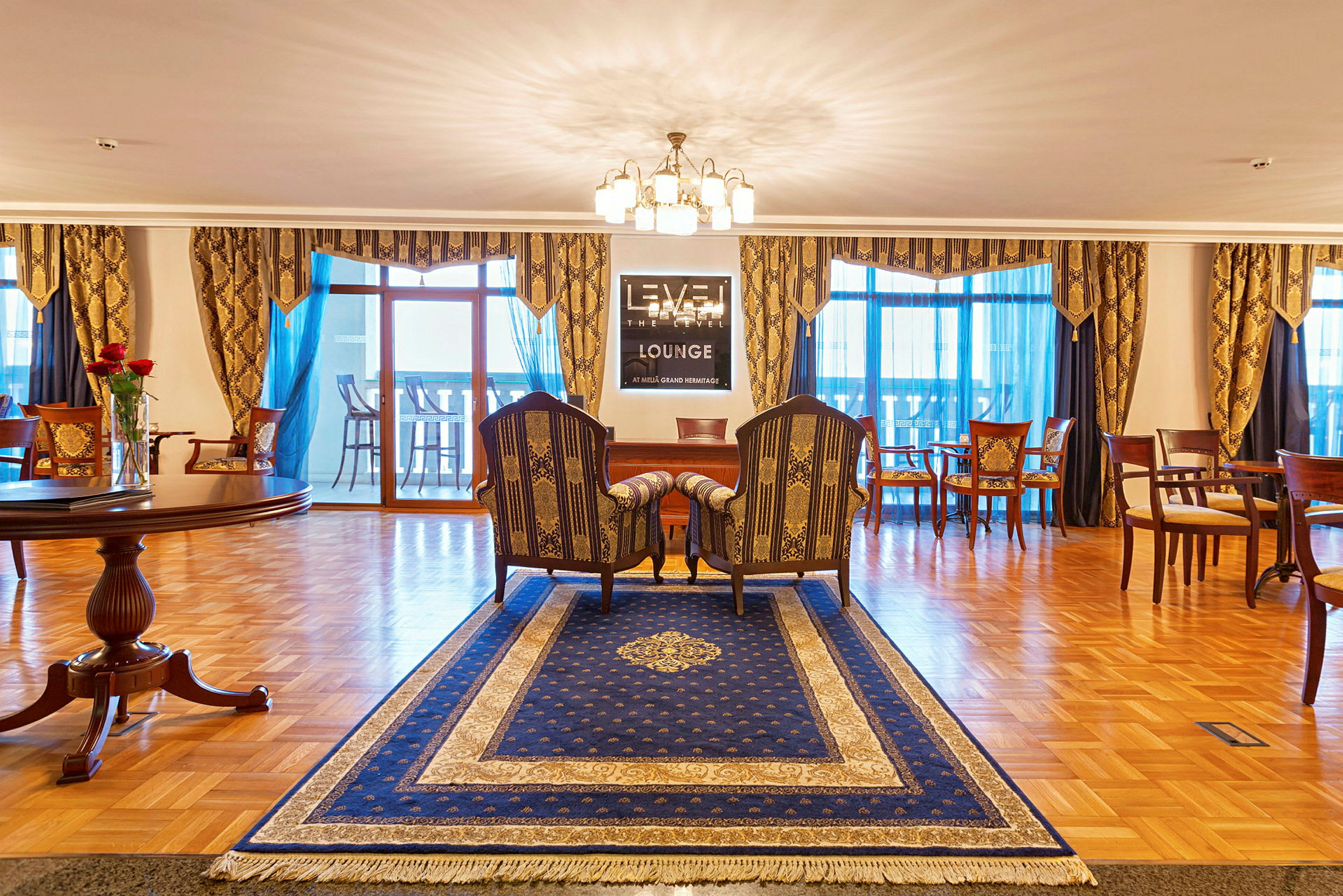 En väl inredd lounge med ett blått mattan, par av stolar och en bord, lampor och utsmyckade gardiner. (AI-genererad text)
