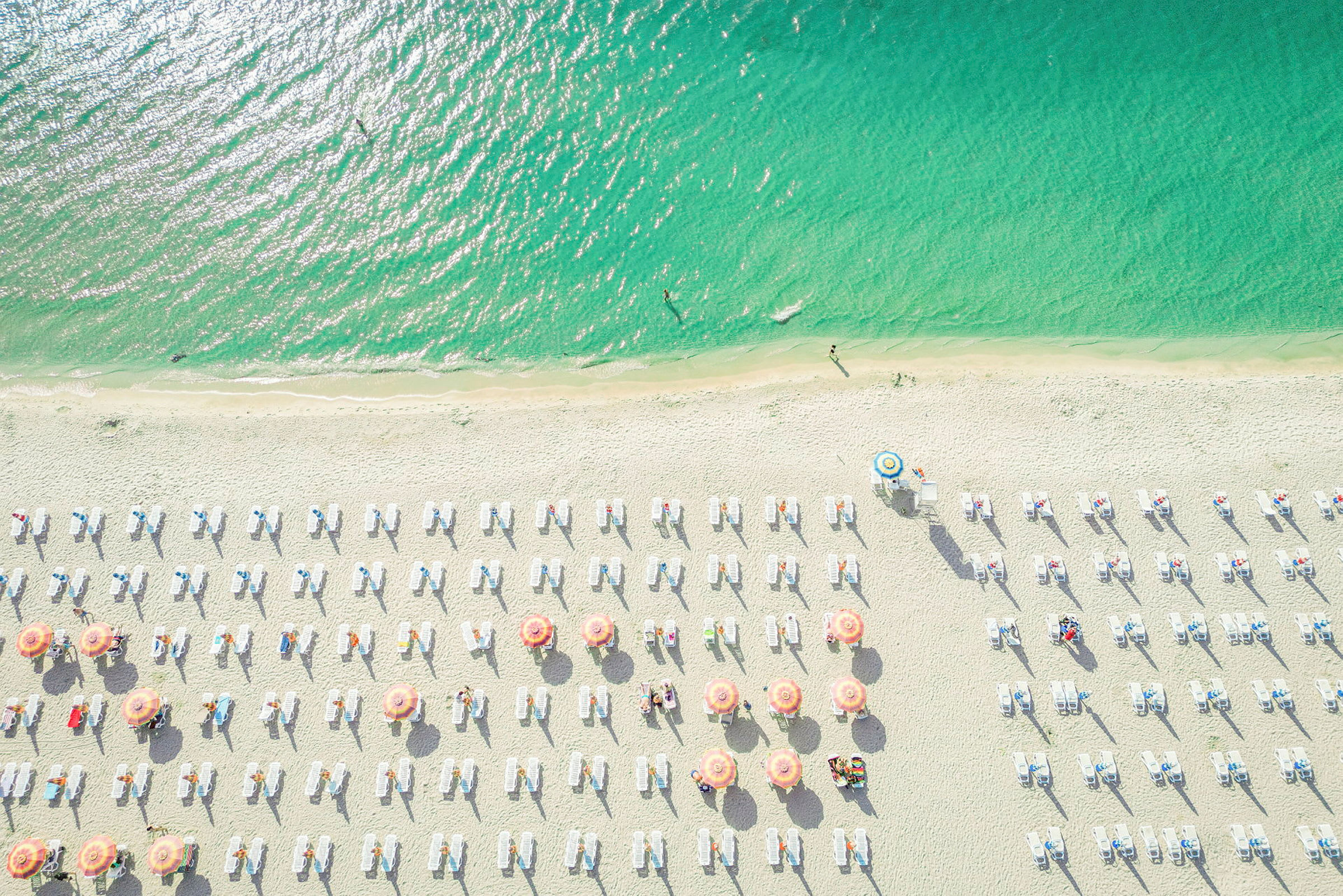 Flygfoto av en strand med parasoller och solstolar. Havet är färgstarkt grönblått. (AI-genererad text)