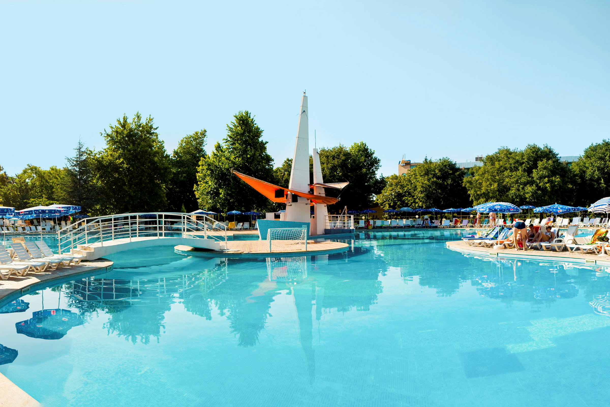Öppen pool med bro och dekorativ båt mitt i, omgiven av solstolar och parasoller. (AI-genererad text)