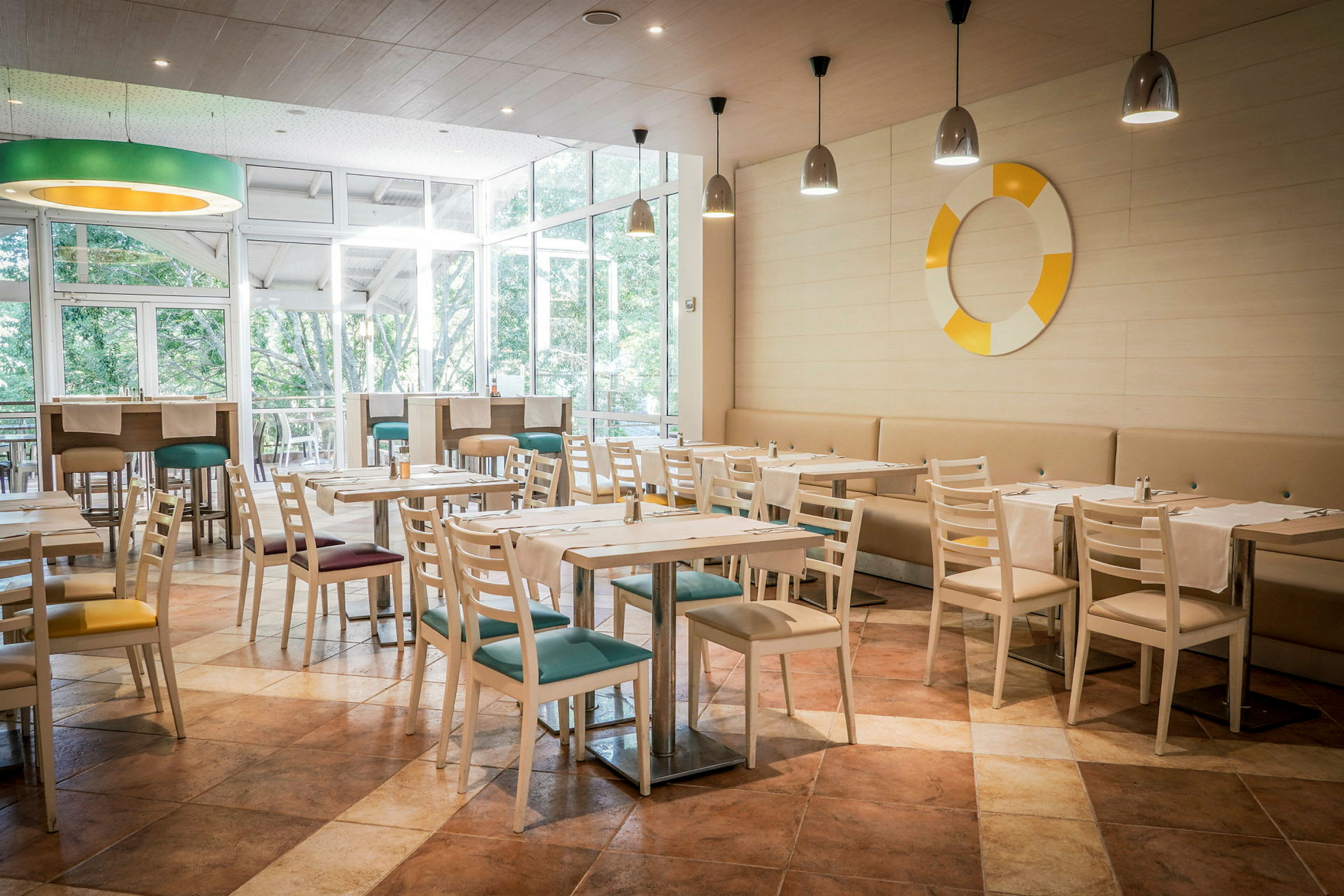 En restaurang med ljusa trästolar och bord, färgglada sittdynor och fönster mot grönska. (AI-genererad text)