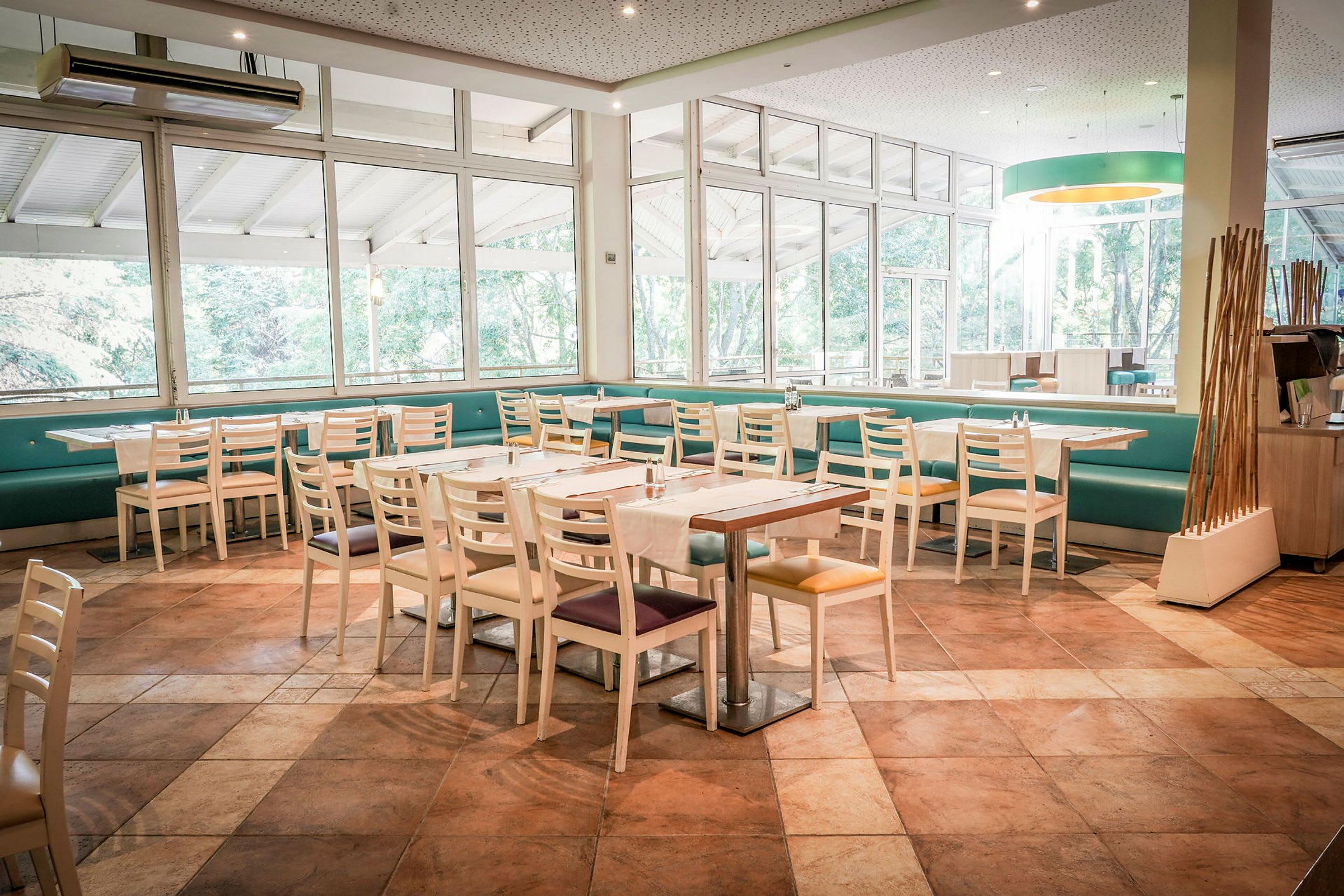 Ett ljust restaurangområde med bord och stolar samt stora fönster som vetter mot grönska. (AI-genererad text)