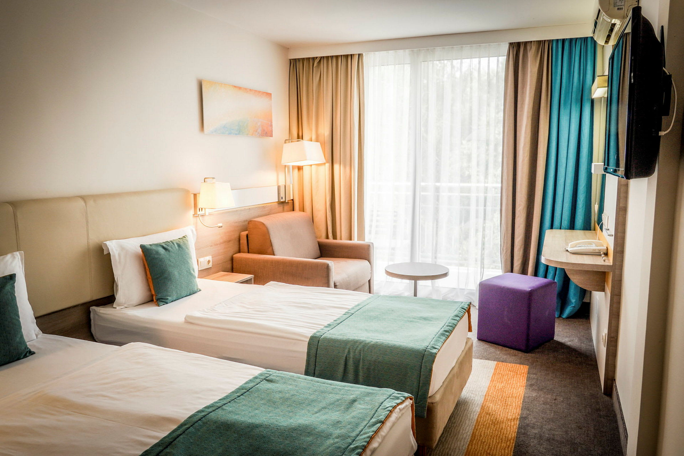 Två enkelsängar med turkosa överkast i ett hotellrum med fönster och sittgrupp. (AI-genererad text)