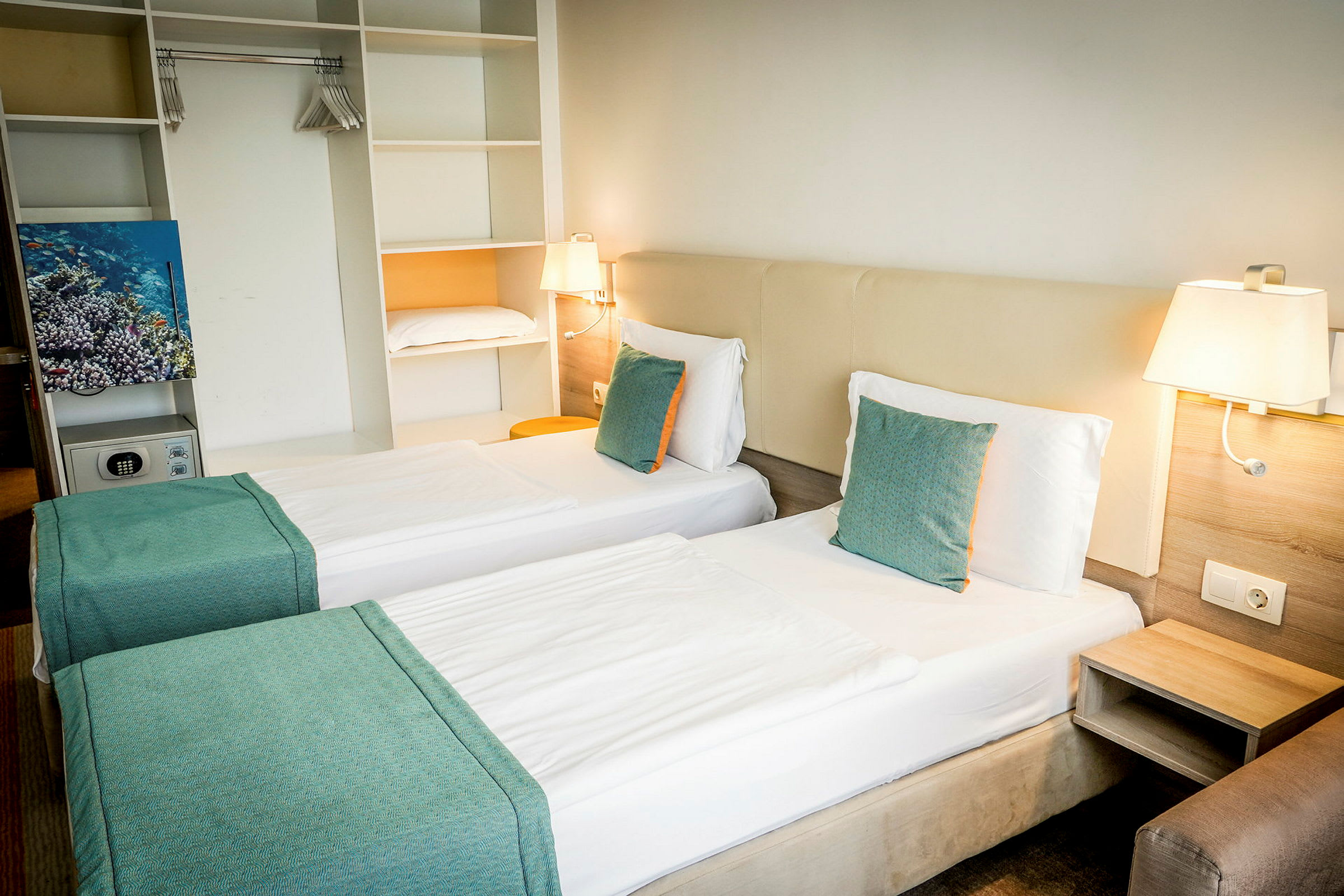 Hotellrum med två enkelsängar, vita lakan och turkosa kuddar. Inbyggd förvaring och vägglampor syns. (AI-genererad text)