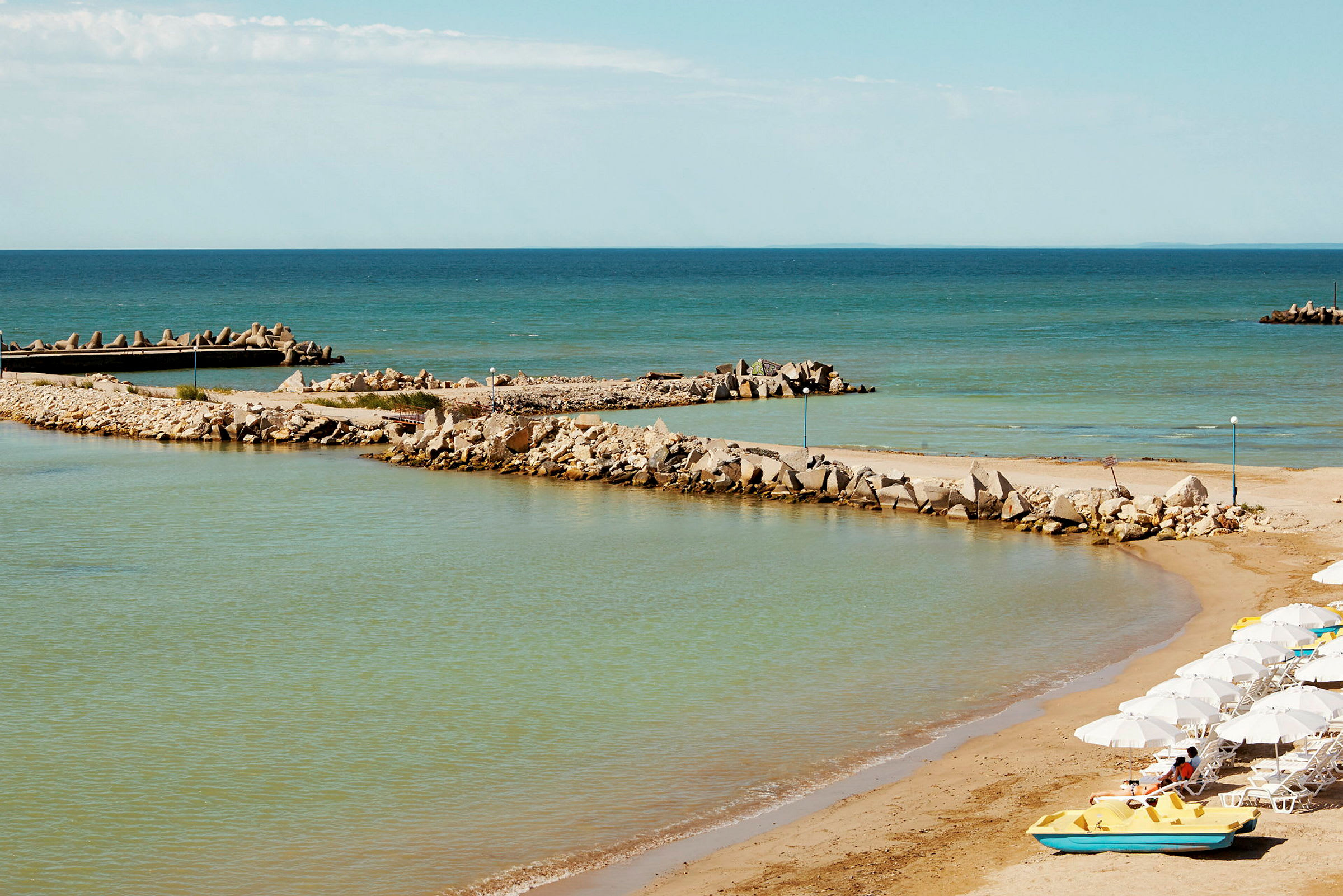 Strand med sand, rad med solsängar och parasoller, vatten i förgrunden och vågbrytare av sten i bakgrunden. (AI-genererad text)