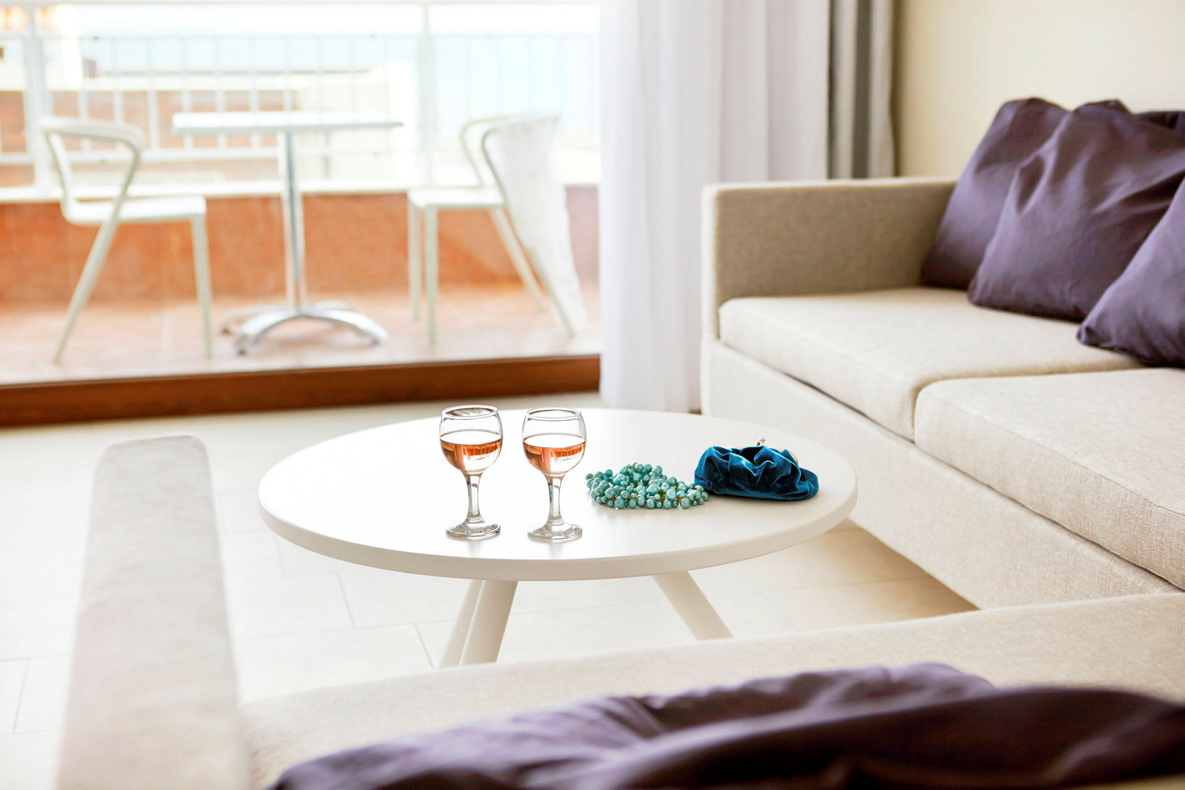 Ett ljust vardagsrum med en vit soffbord som har två vinglas, ett blått smyckeset eller påse och pärlor. En beige soffa med dekorativa lila kuddar bredvid, och en balkong med bord och stolar synlig utanför glasfönstret. (AI-genererad text)