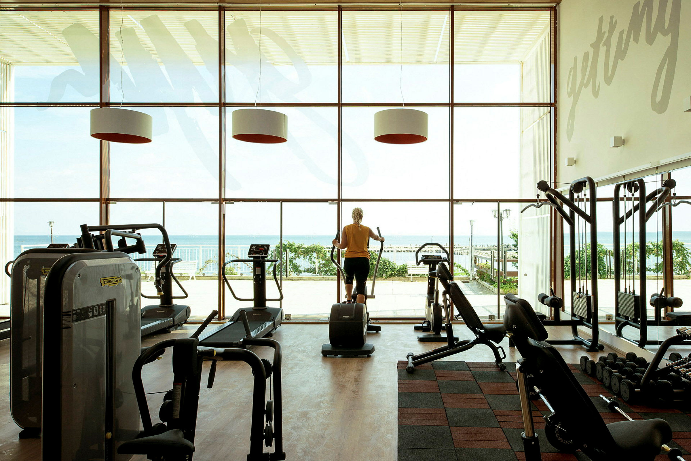 Ett gym med träningsmaskiner framför stora glasfönster med utsikt över havet. (AI-genererad text)