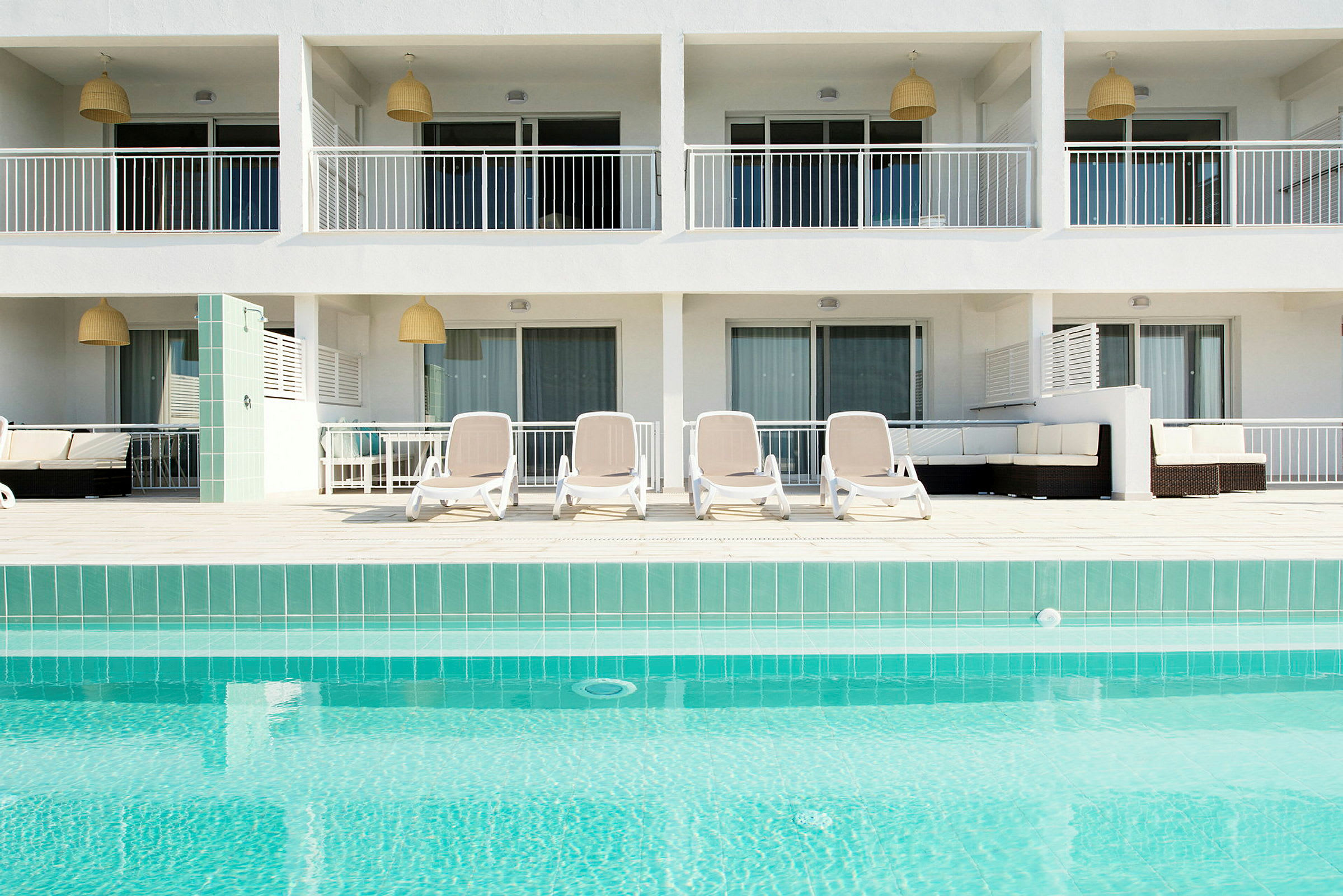 En pool med turkost vatten framför en byggnad med balkonger och solstolar på en terrass. (AI-genererad text)