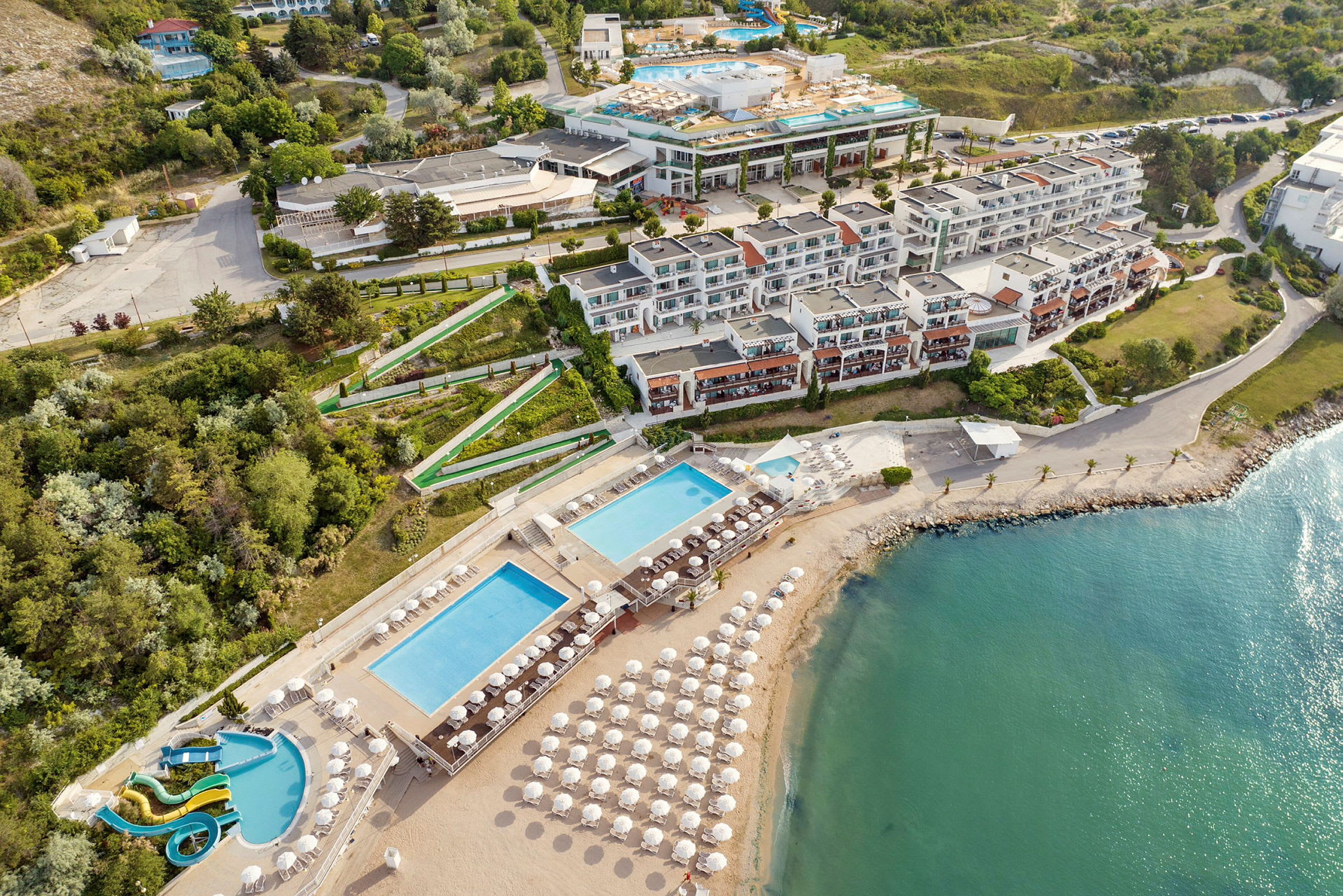 Bild av en resort med byggnader, pooler och en strand med solstolar vid havet. (AI-genererad text)