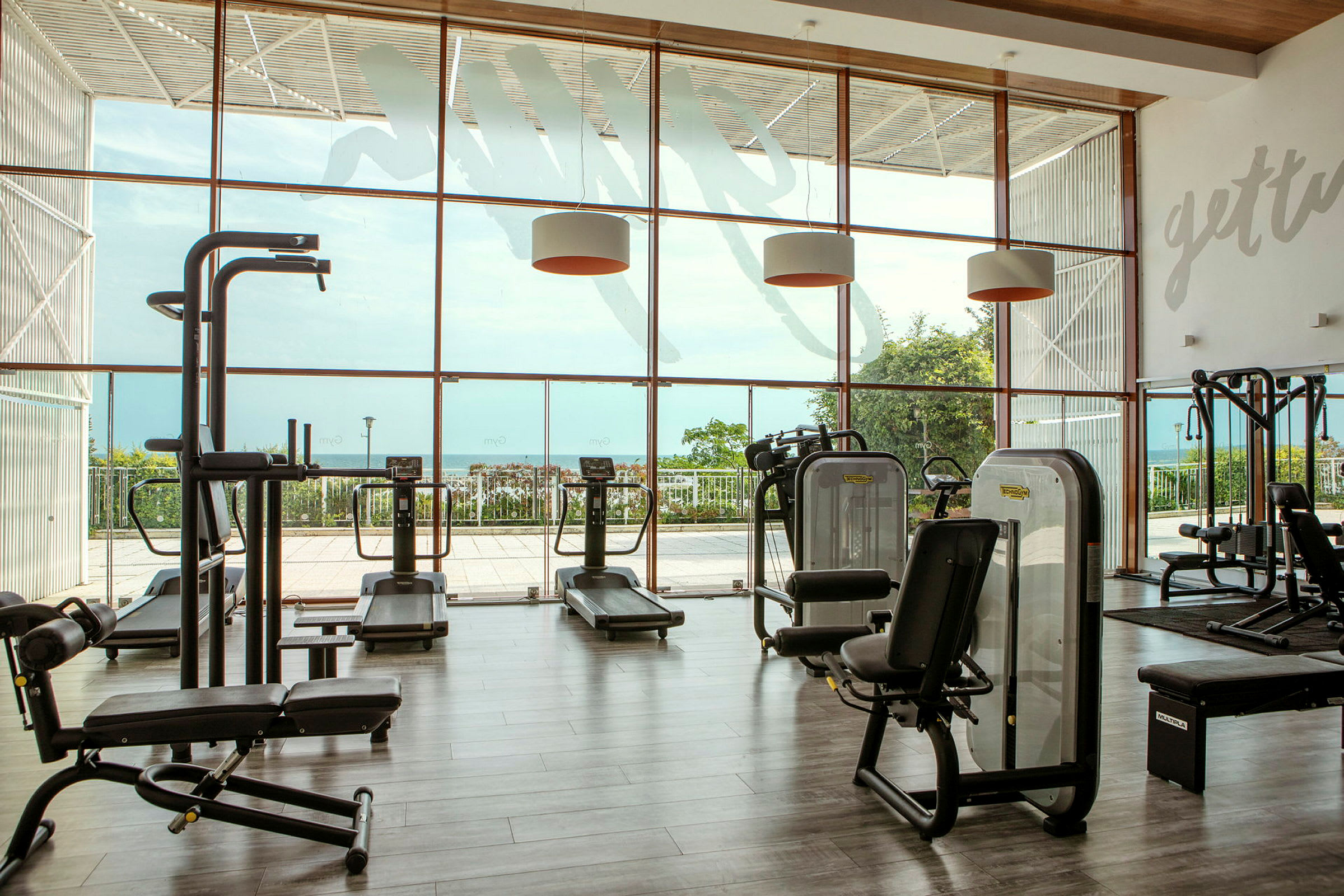 Ett gym med träningsmaskiner och stora fönster med utsikt över ett grönområde och havet. (AI-genererad text)