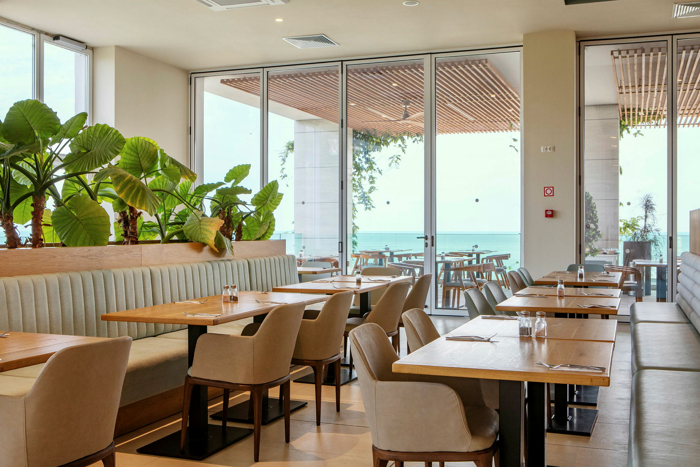 En restaurang med bord, stolar och växter vid stora fönster med utsikt över havet. (AI-genererad text)