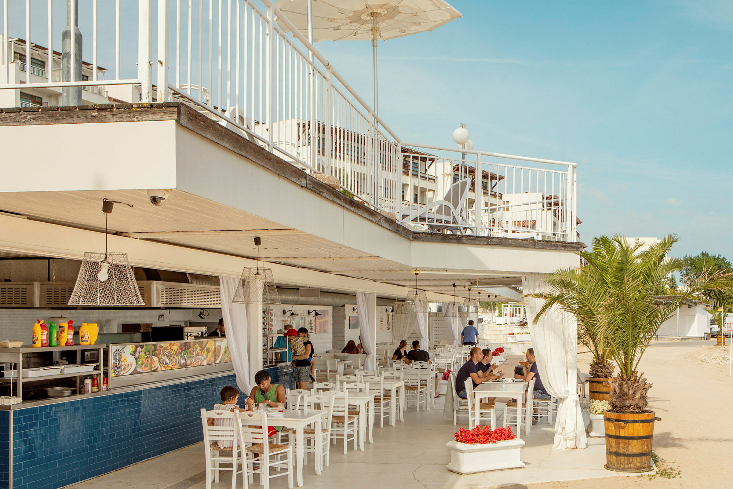 En utomhusrestaurang med vita möbler, hängande lampor och en takterrass. Människor sitter och äter under taket. En palm är också synlig bredvid restaurangen. (AI-genererad text)