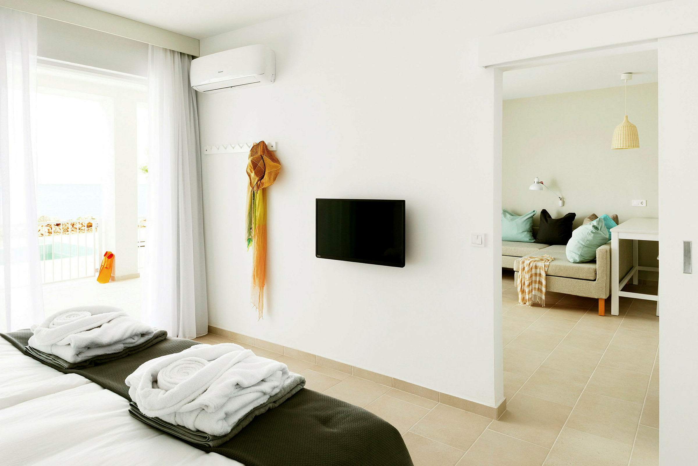 Ett ljust hotellrum med en bäddad säng, en väggmonterad TV och en sittgrupp. Balkong med utsikt syns i bakgrunden. (AI-genererad text)
