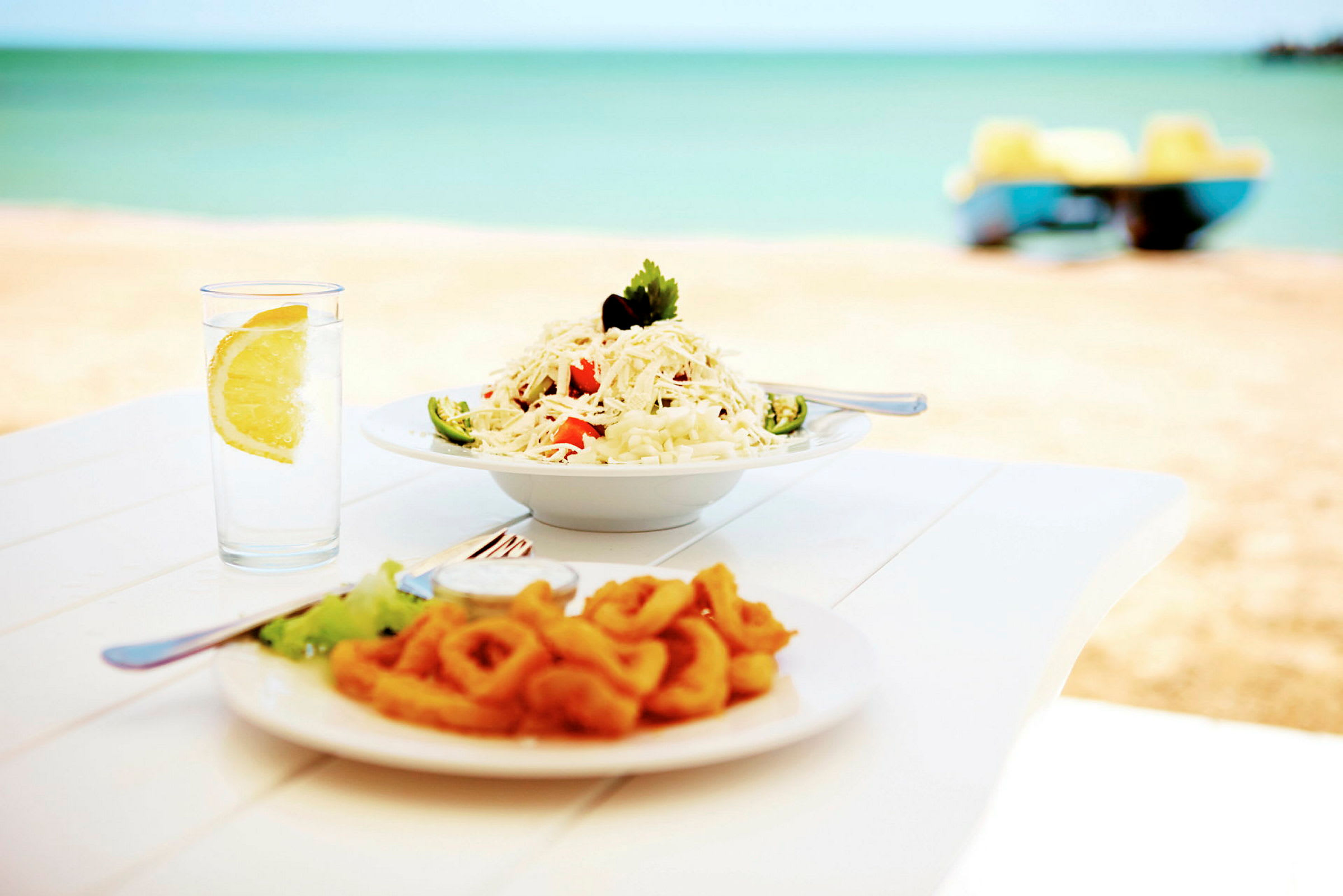 En tallrik med pasta och en tallrik med friterade bläckfiskringar serverade vid ett bord vid stranden, med ett glas vatten och en citronklyfta bredvid. (AI-genererad text)