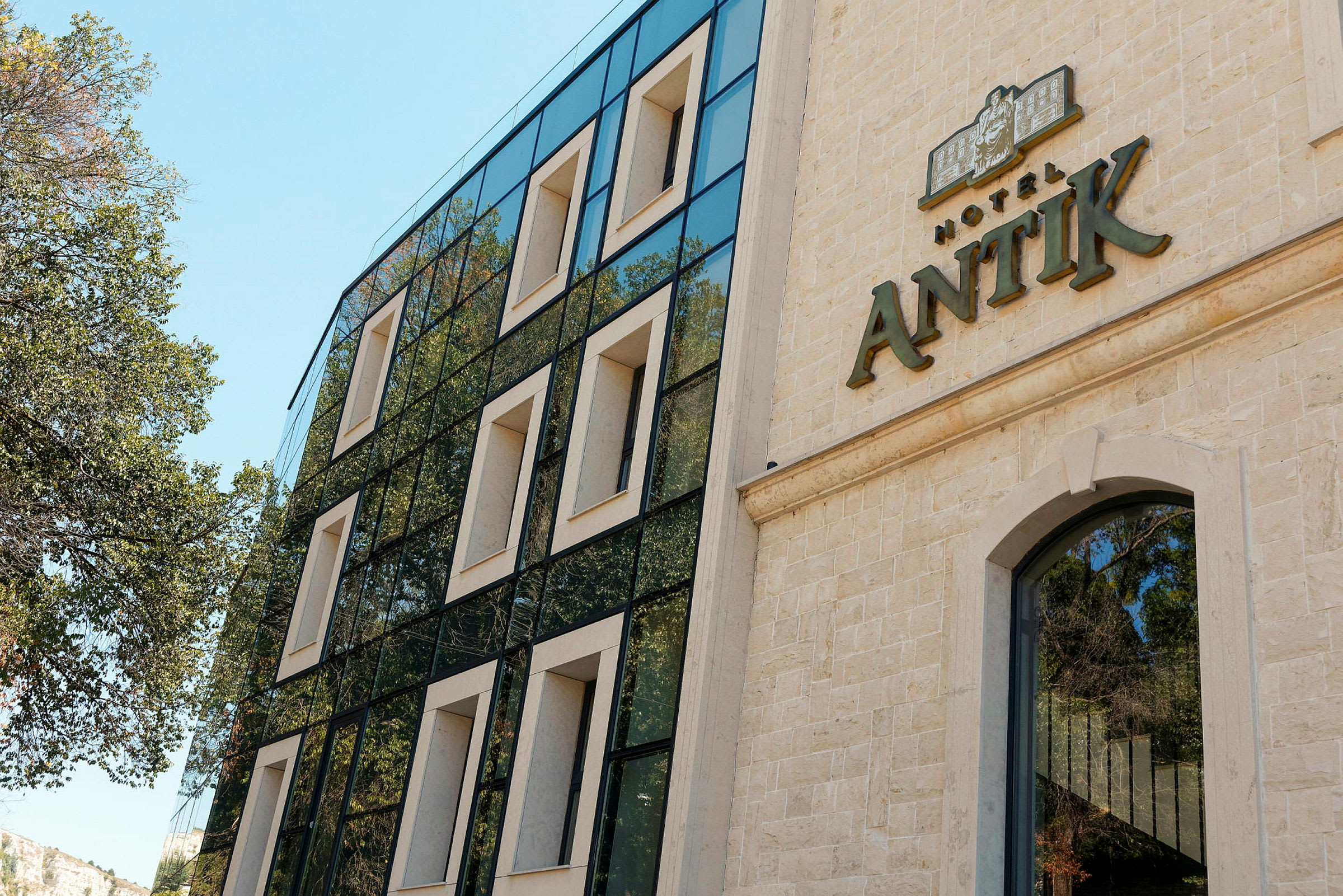 Fasad av ett hotell med namnet "Hotel Antik" synligt på byggnaden. Den moderna byggnaden har stora fönster och omges av träd. (AI-genererad text)