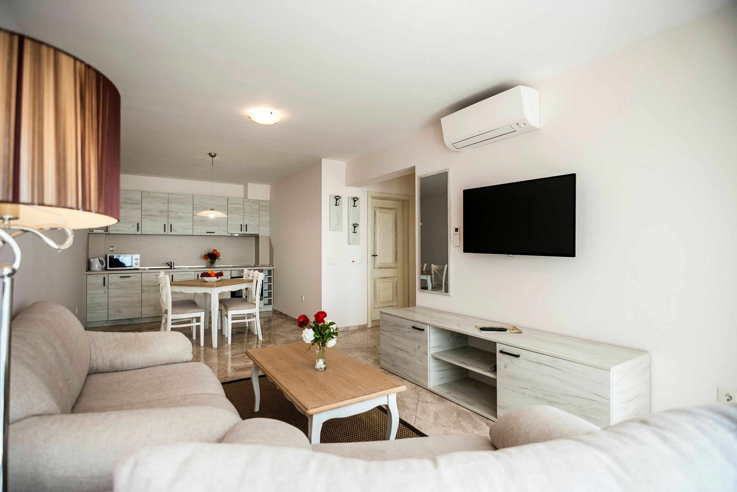En modern vardagsrum med en grå soffa, ett träbord och en väggmonterad TV. Köket i bakgrunden har ljusa skåpluckor och en matplats. (AI-genererad text)