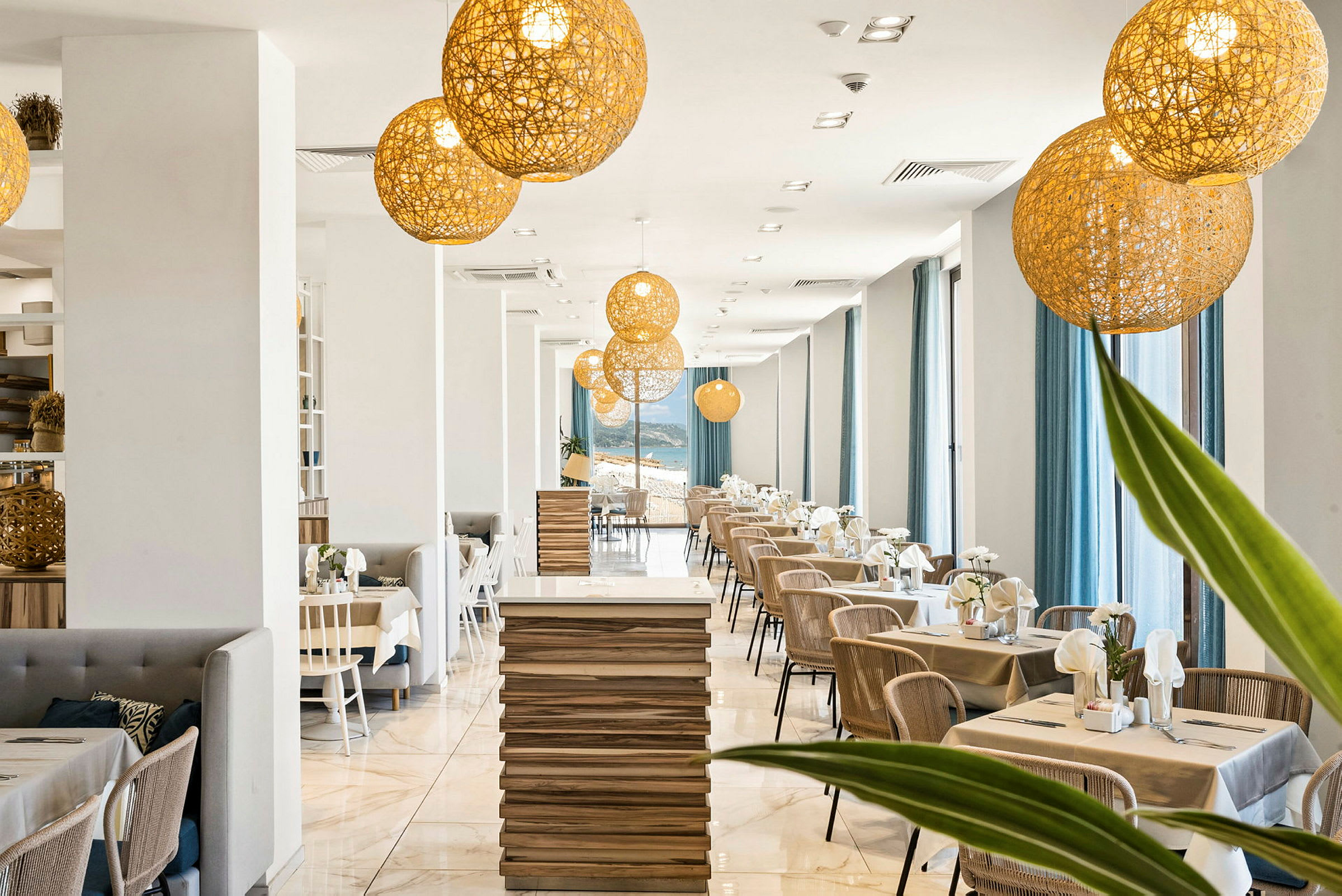En modern restaurang med en ljus inredning innehållande träbord, stolar och stora fönster med blåa gardiner. Dekorativa belysningsarmaturer hänger från taket. (AI-genererad text)