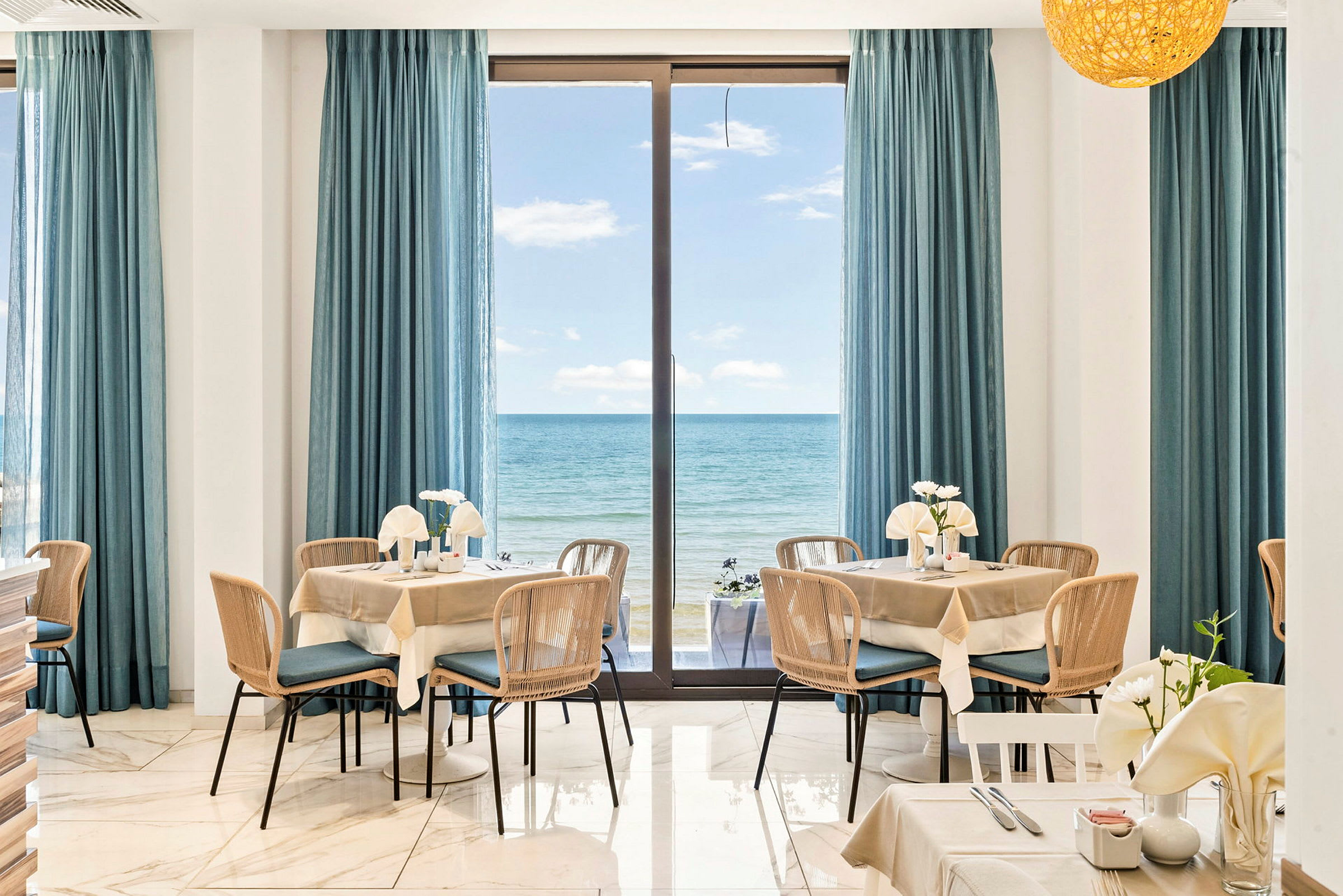 Interiören i en restaurang med bord och stolar, fönster täckta med blå gardiner, och utsikt över havet. (AI-genererad text)