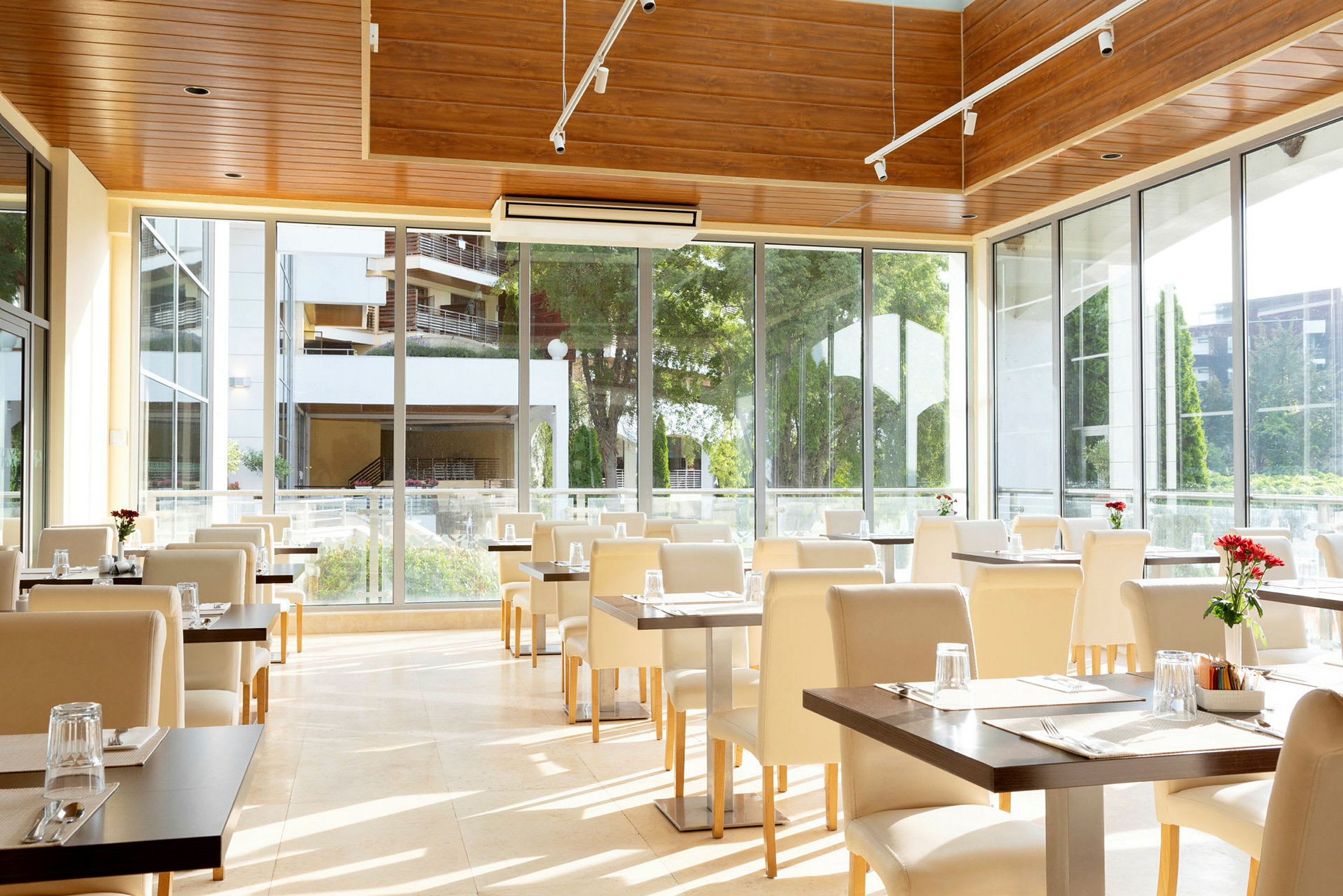 Restaurang med stora fönster, vita stolar och dukade bord med glas och röda blommor. (AI-genererad text)