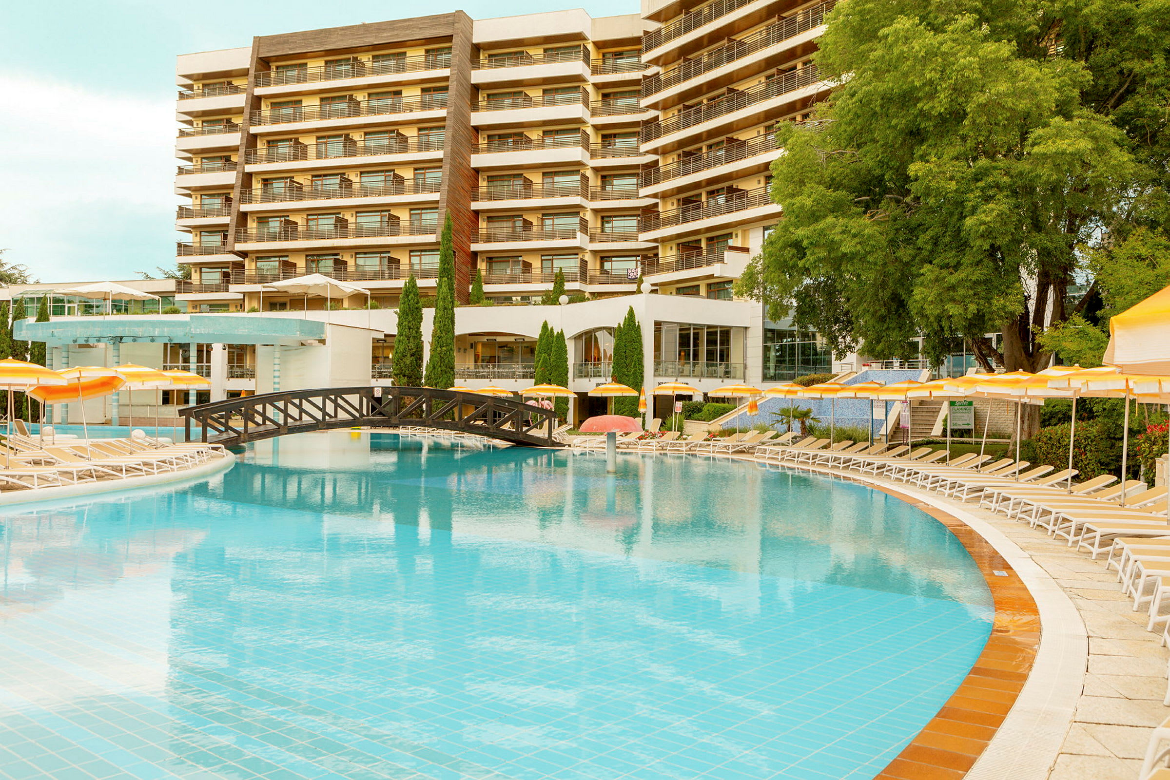 Hotellfasad med flera våningar synlig i bakgrunden bakom en stor pool och solstolar. Utsmyckade balkonger och grönska omger området. (AI-genererad text)