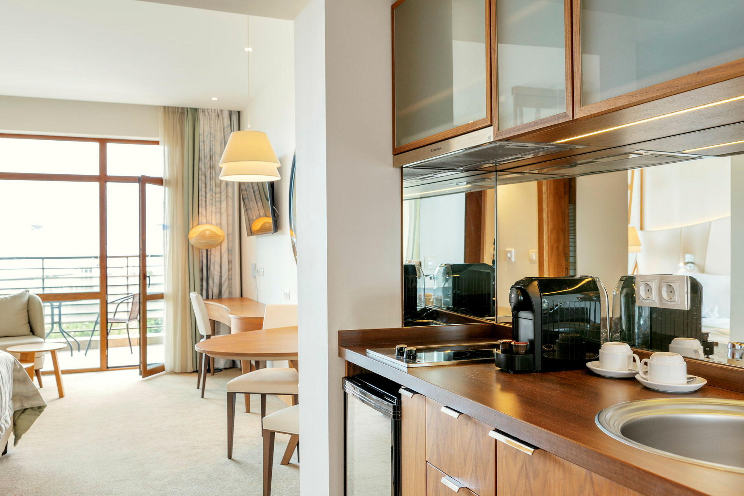 Modernt hotellrum med balkong, bord, stolar, kaffebryggare och köksyta. (AI-genererad text)
