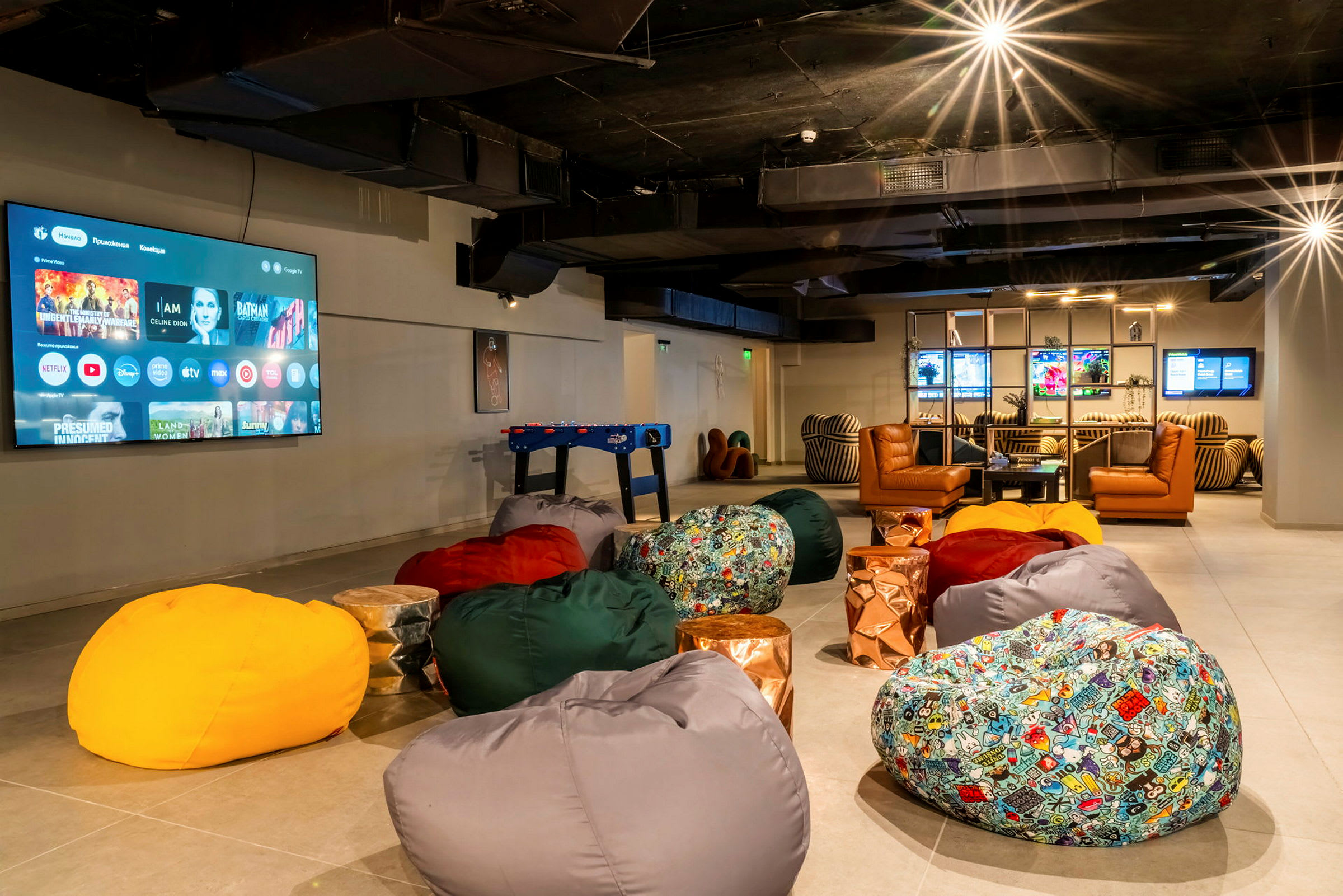 Ett rum med stora sittpuffar, en stor TV-skärm på väggen, och en avkopplingshörna med skinnfåtöljer. (AI-genererad text)