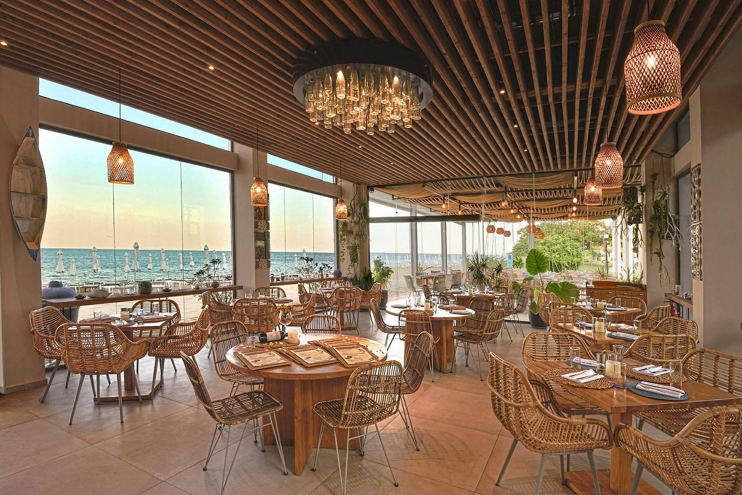 Restaurang med träbord och rottingstolar, stora fönster visar vy mot havet. (AI-genererad text)
