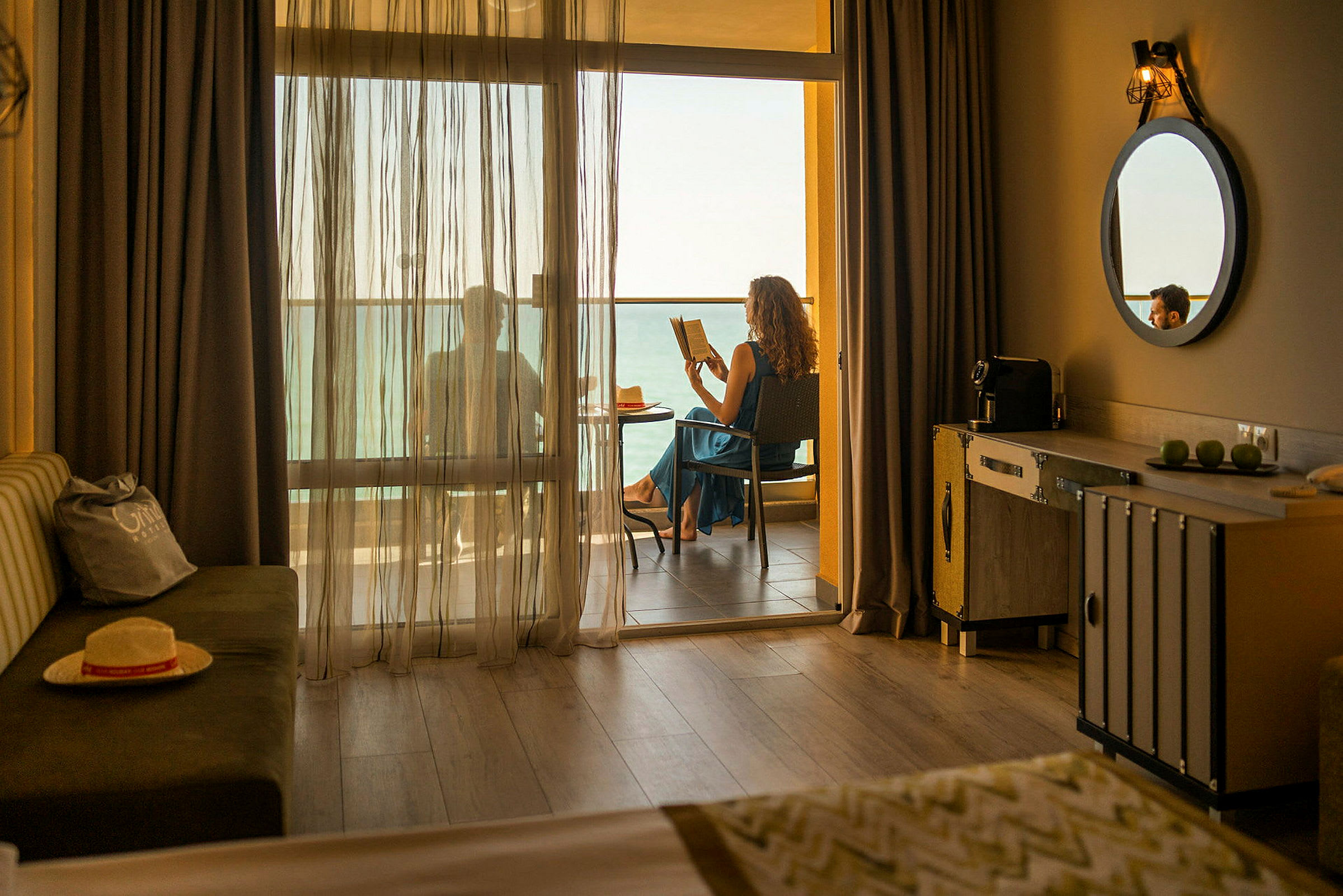 Hotellrum med en balkong där någon sitter och läser med havsutsikt. (AI-genererad text)