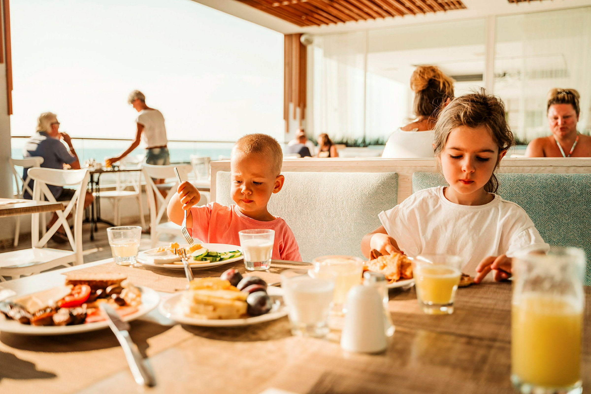 Barn sitter vid ett bord och äter medan människor syns i bakgrunden på en restaurang med utsikt. (AI-genererad text)